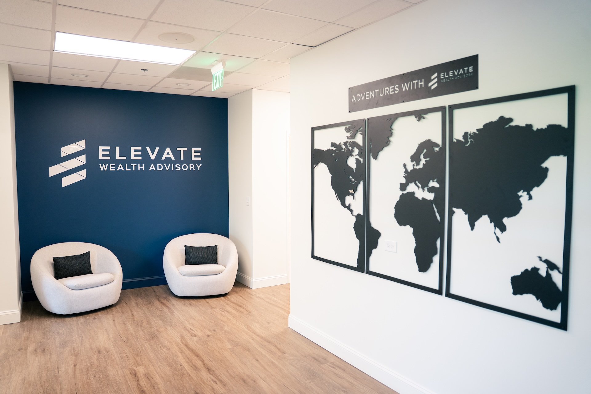 Office interior with two white chairs, black pillows, blue wall with 'ELEVATE WEALTH ADVISORY' logo, and a wall display of three world maps with the text 'ADVENTURES WITH ELEVATE'.