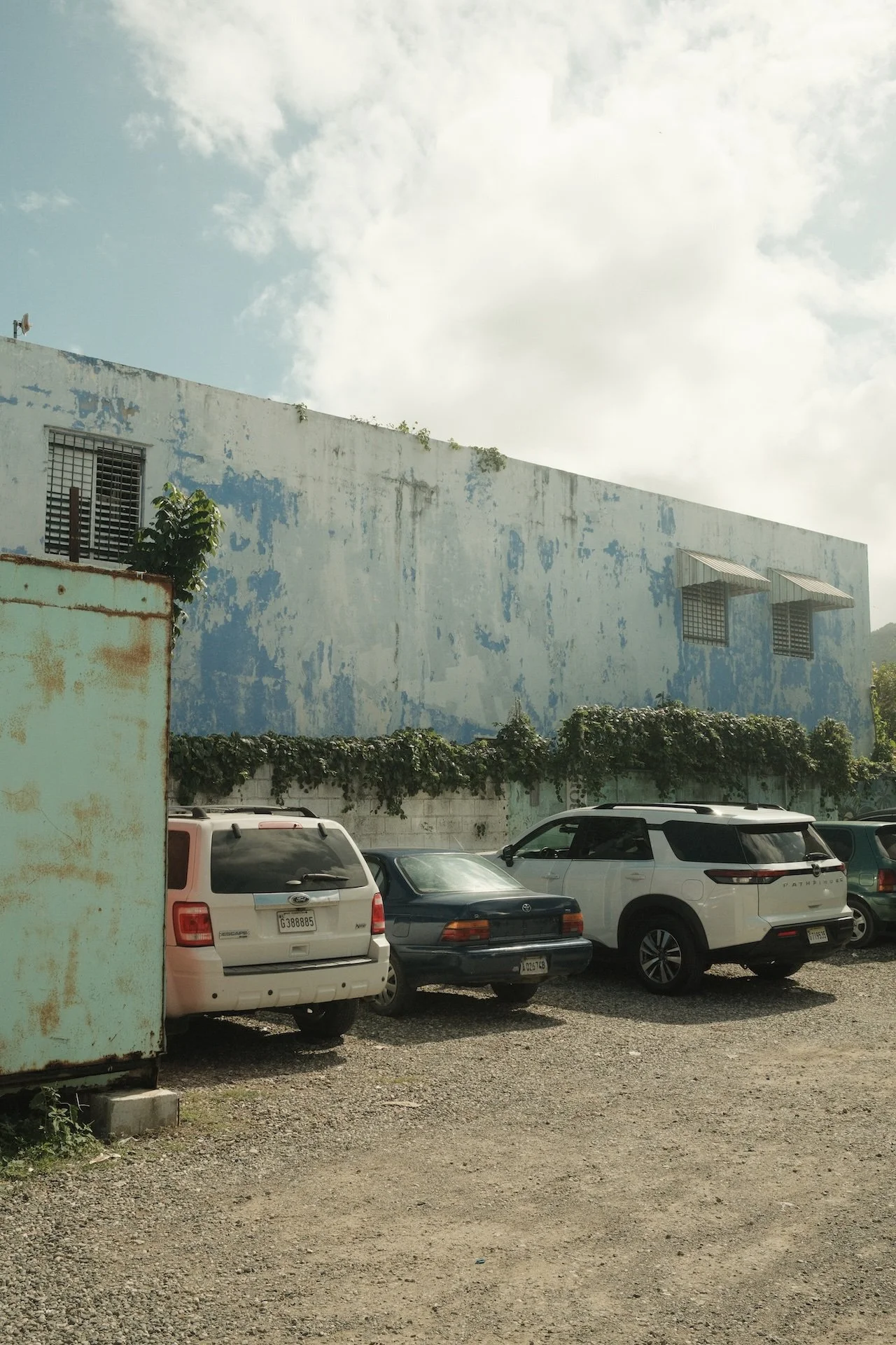 A parking lot with several cars parked in front of a weathered blue warehouse with small windows and awnings, under a partly cloudy sky.