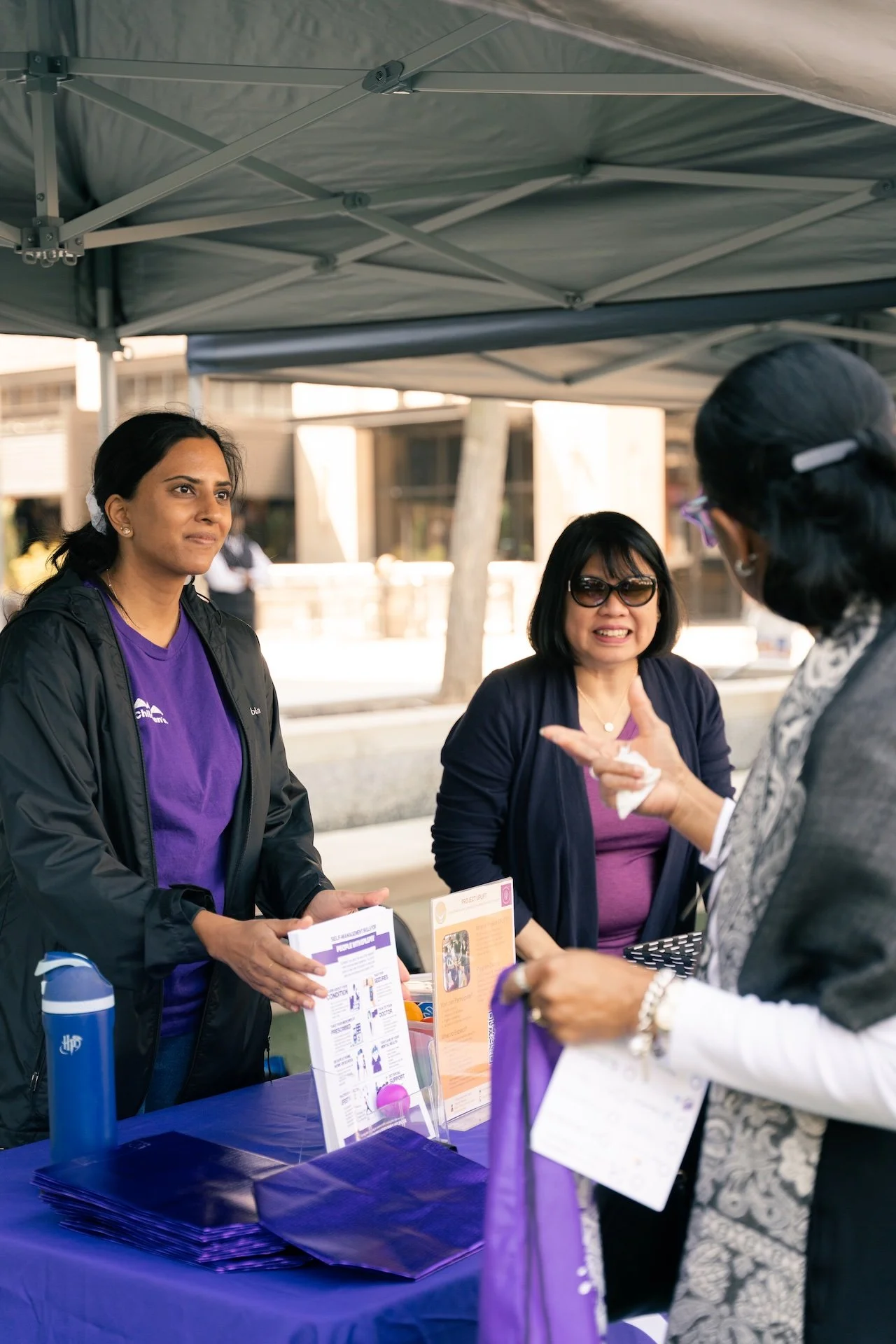 Three women at a booth under a canopy, engaging in conversation. The woman on the left holds pamphlets, the woman in the middle wears sunglasses, and the woman on the right is speaking, all participating in a discussion at an outdoor event.