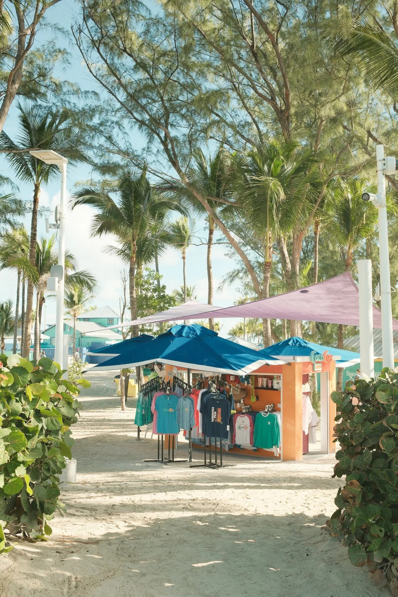 A small outdoor souvenir shop on a sandy beach, shaded by large blue and pink umbrellas, with colorful T-shirts hanging on racks, surrounded by lush green palm trees and tropical vegetation, under a clear blue sky.