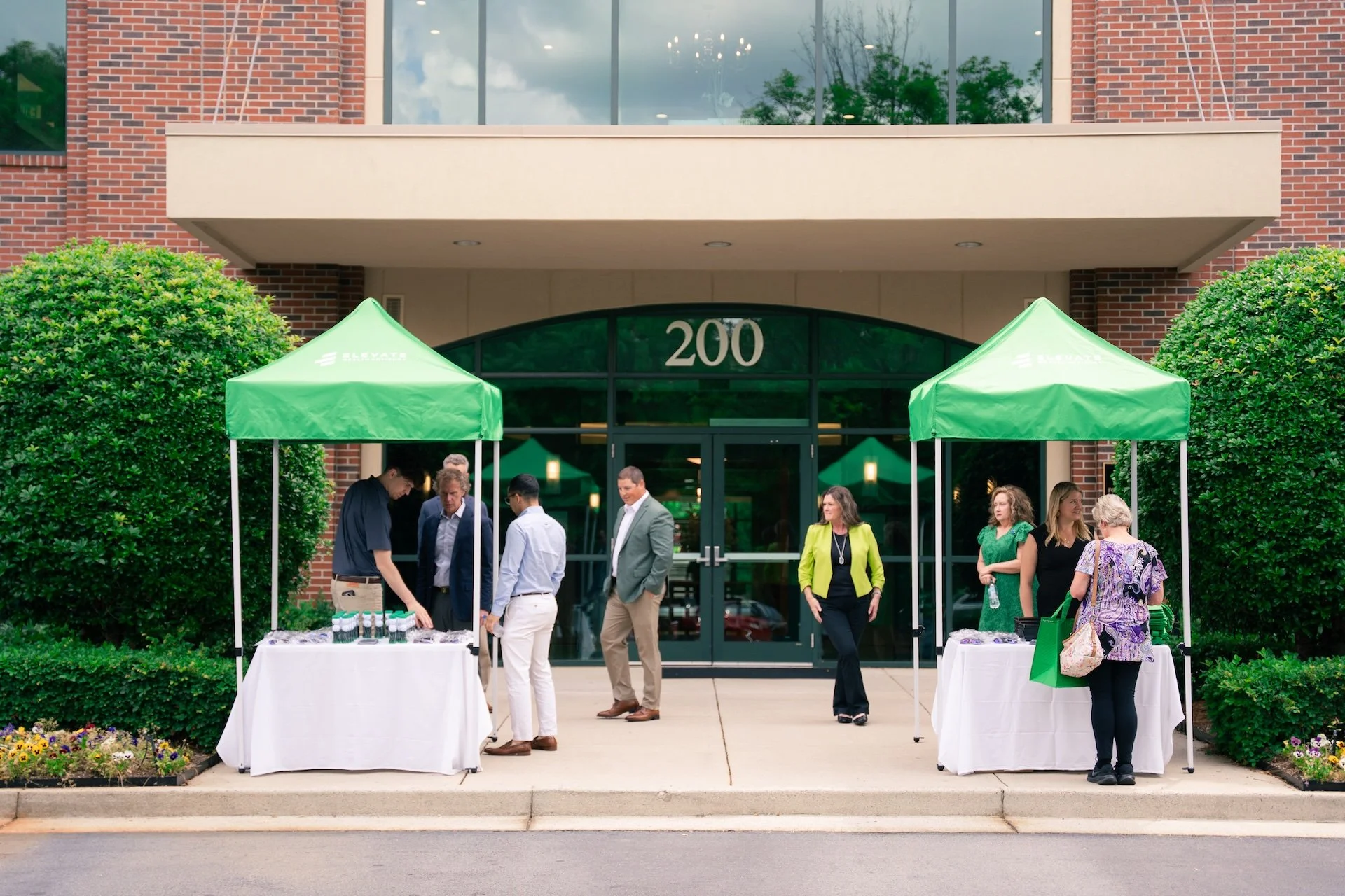People gathered outside a building with the number 200 above the door, under green tents, engaging in a registration or event activity.