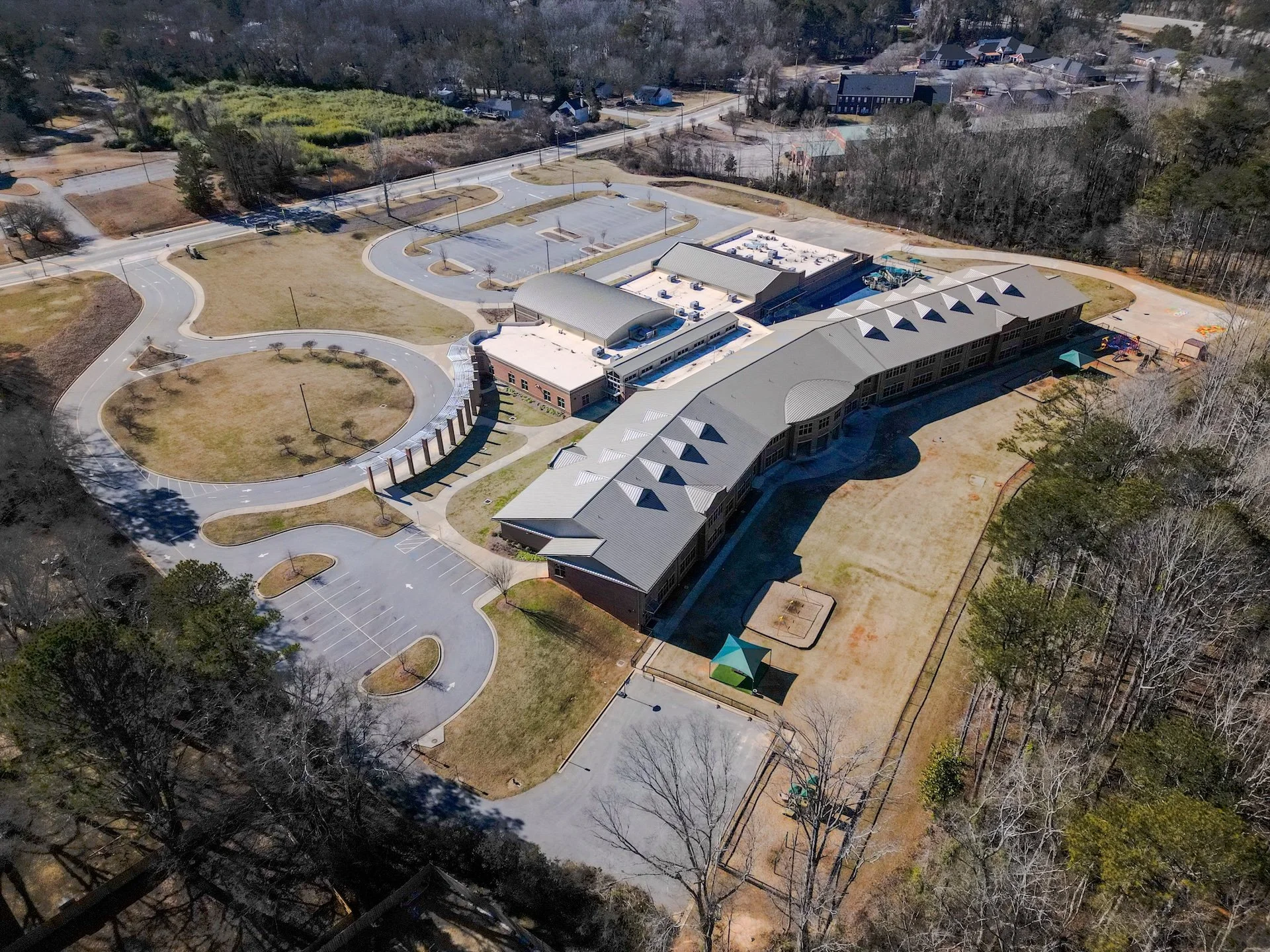 Aerial view of a school campus with a large building, parking lots, playground, and surrounding trees.
