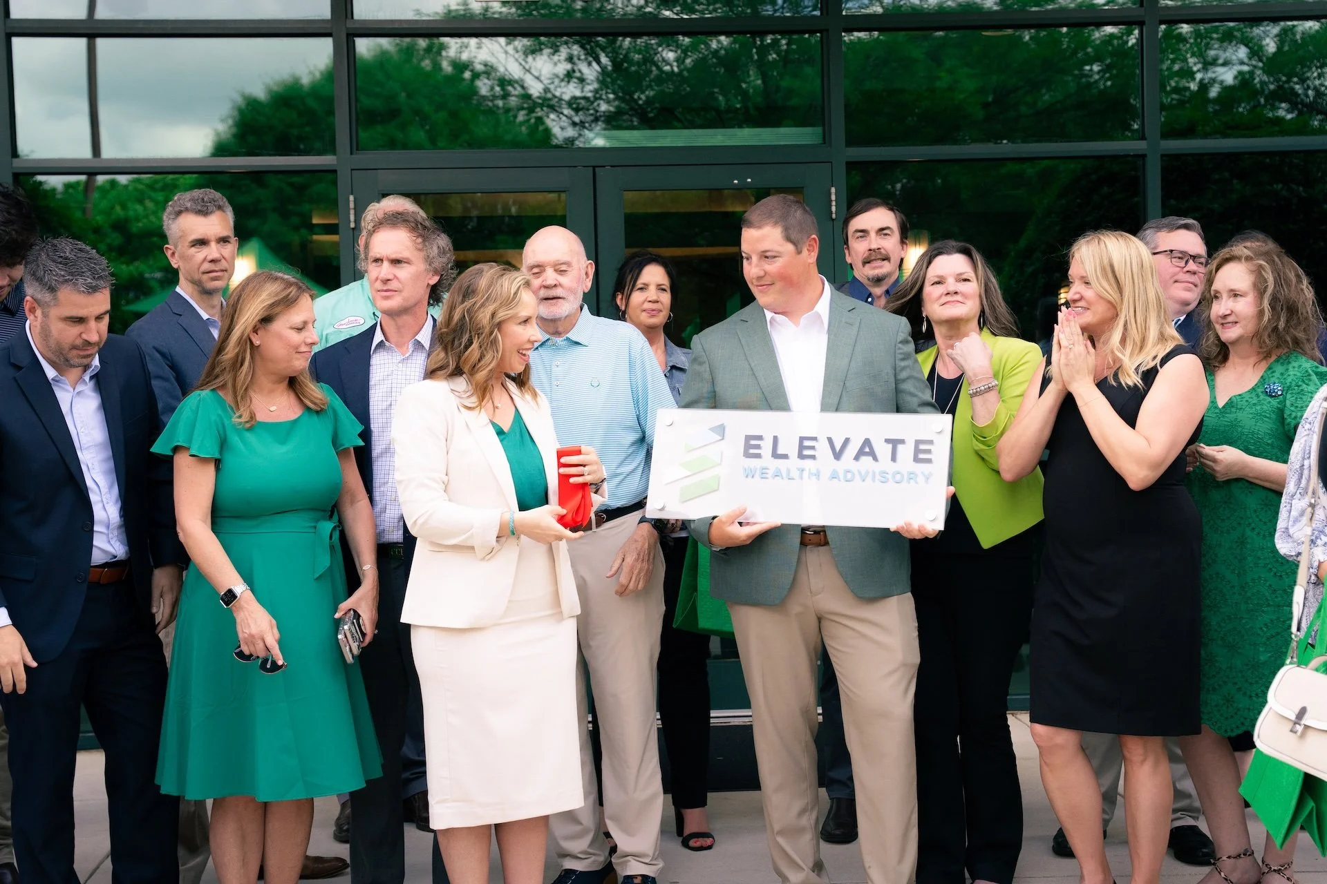 A group of professionally dressed people gathered outside a modern glass building for a presentation or event, with some holding a sign that says 'Elevate Wealth Advisory'.