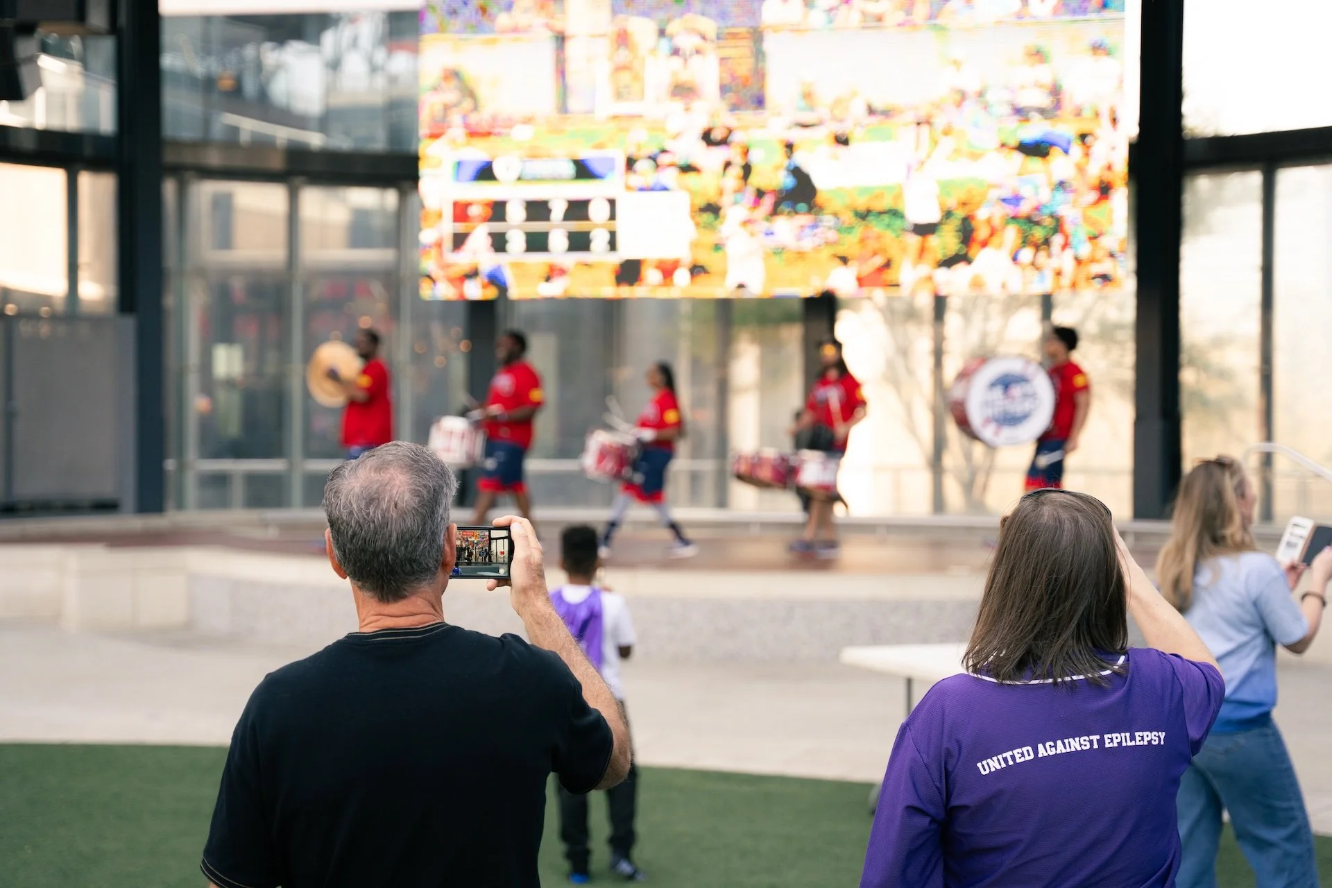 People watching a parade or performance of a marching band on a stage in front of a large colorful digital display, with some individuals taking photos or videos.
