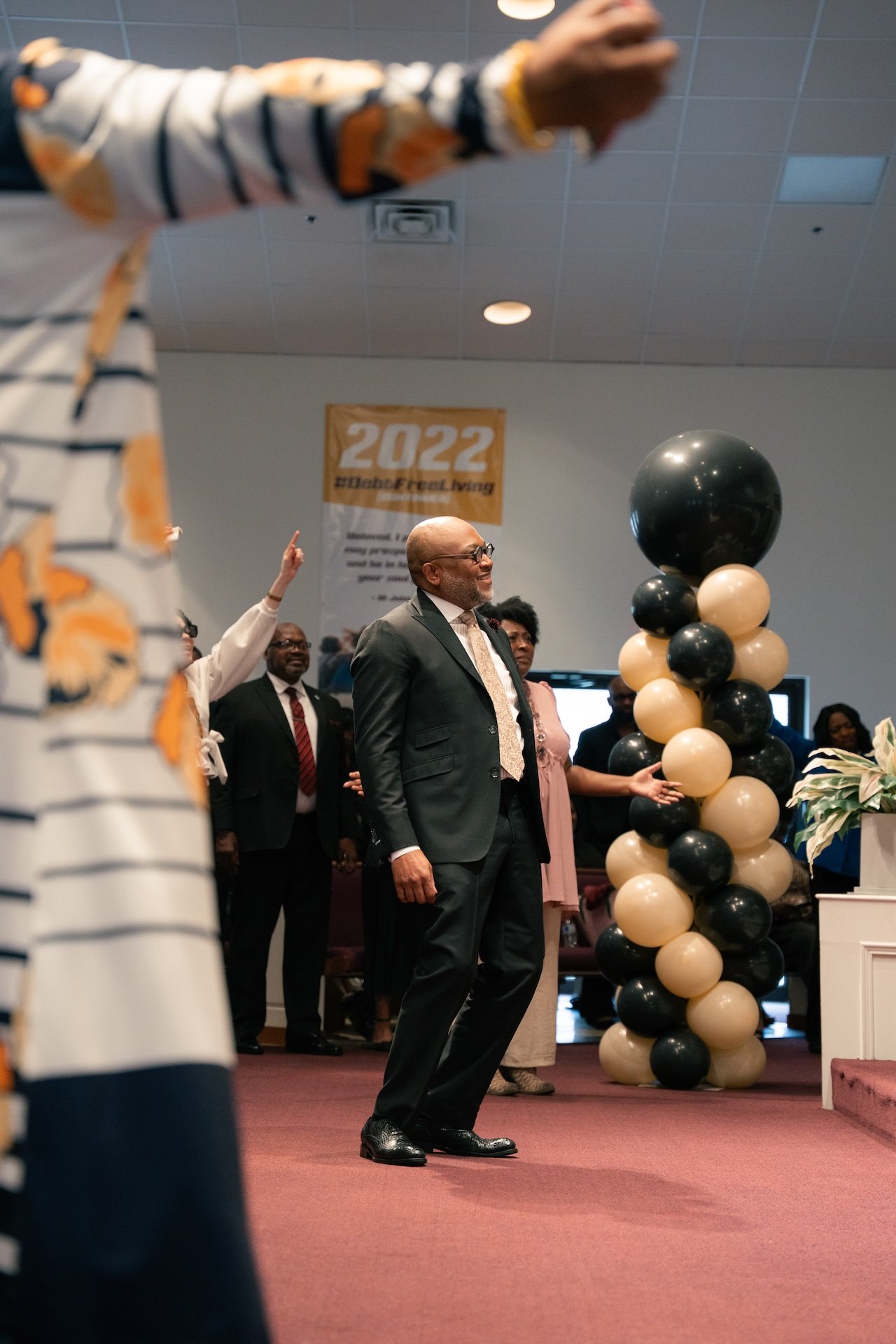 A group of people in formal attire inside a church, with one man standing in the foreground and others raising their hands, along with black and beige balloons, and a banner that reads '2022' in the background.