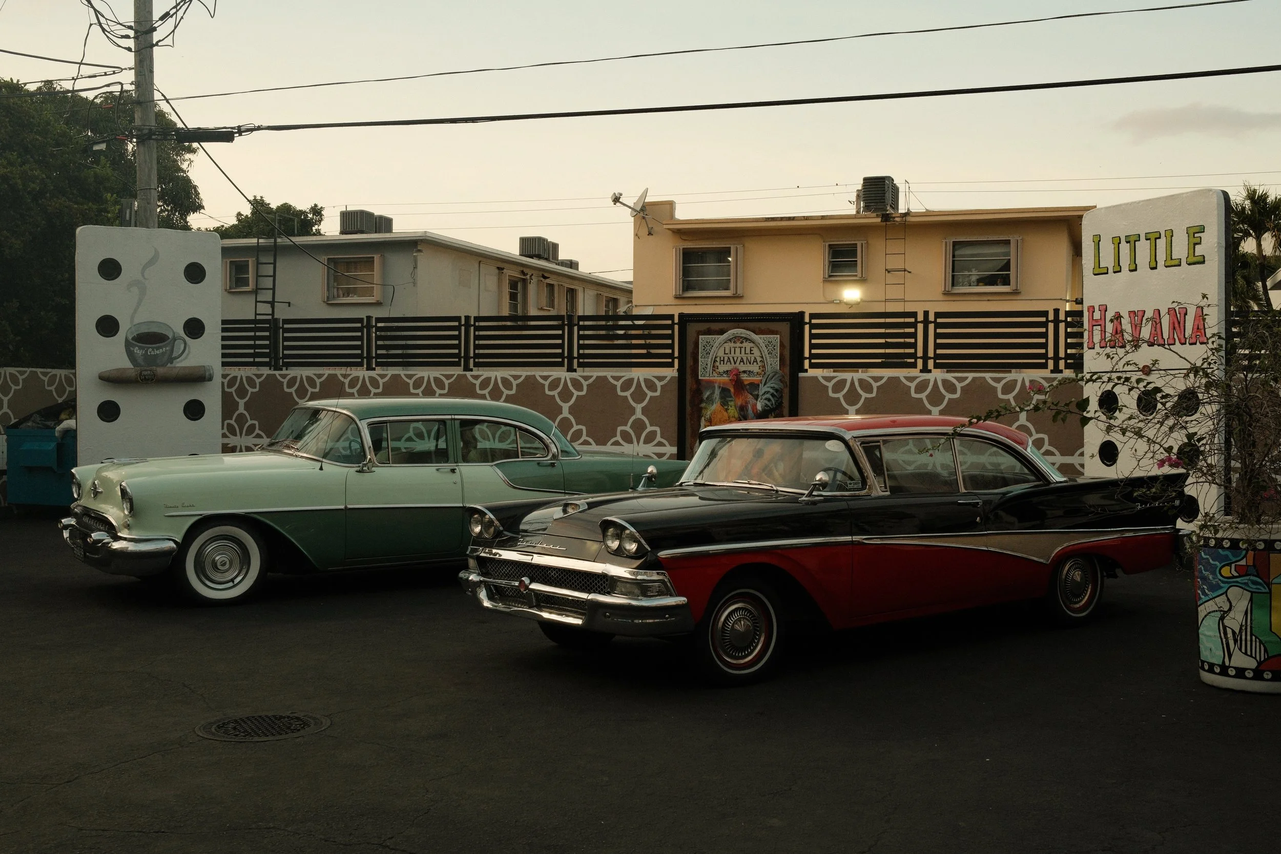 Two vintage 1950s cars parked in front of a mural that reads "Little Havana." One car is green and white, and the other is black and red.