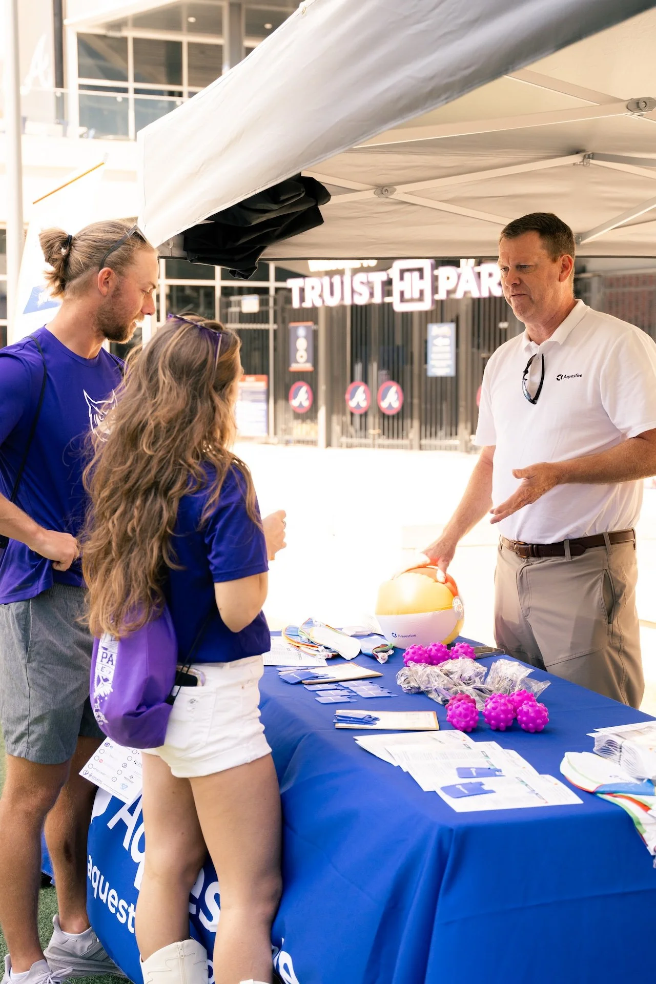 A man in a white polo shirt and khaki pants is explaining something at an outdoor event, with a table of informational materials, pink spiky balls, and a beach ball, while two young adults in blue shirts listen, under a white canopy at Truist Park.