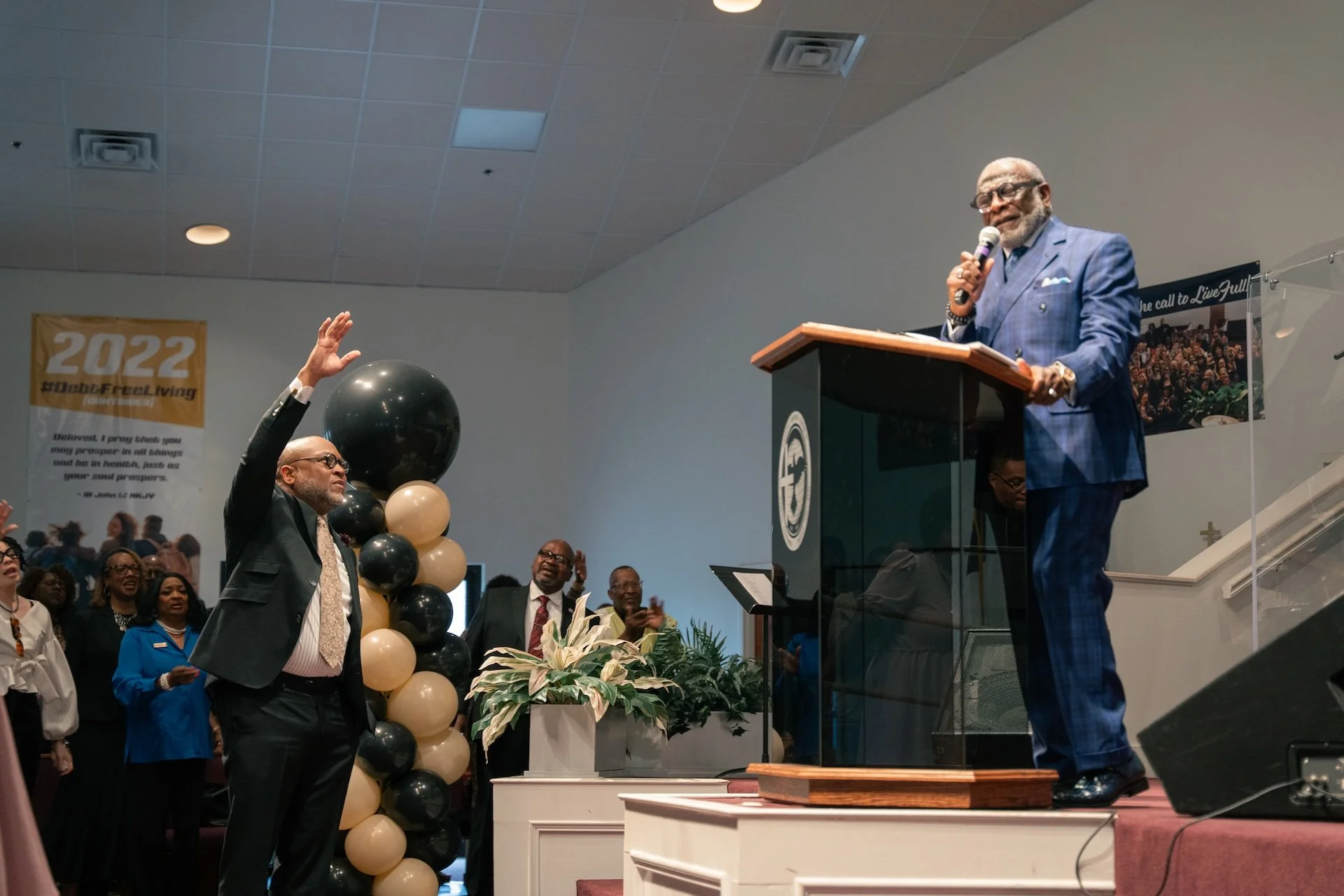 A man in a blue checkered suit speaking at a podium on stage, with people in the congregation raising their hands in response, during a church service or event.