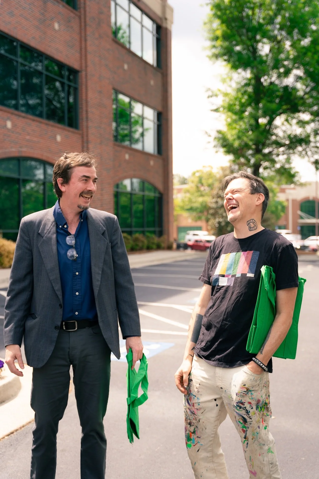 Two men are standing outdoors in a parking lot, laughing and talking to each other. One is dressed in a suit and holding a green cloth, the other is casually dressed with paint-stained pants and a t-shirt. They are near a brick building with large wi