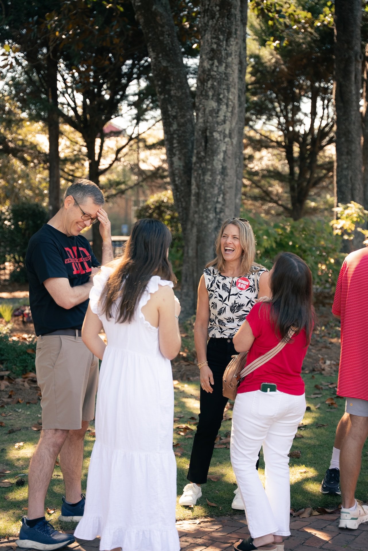 Group of people outdoors, laughing and chatting, surrounded by trees in sunlight