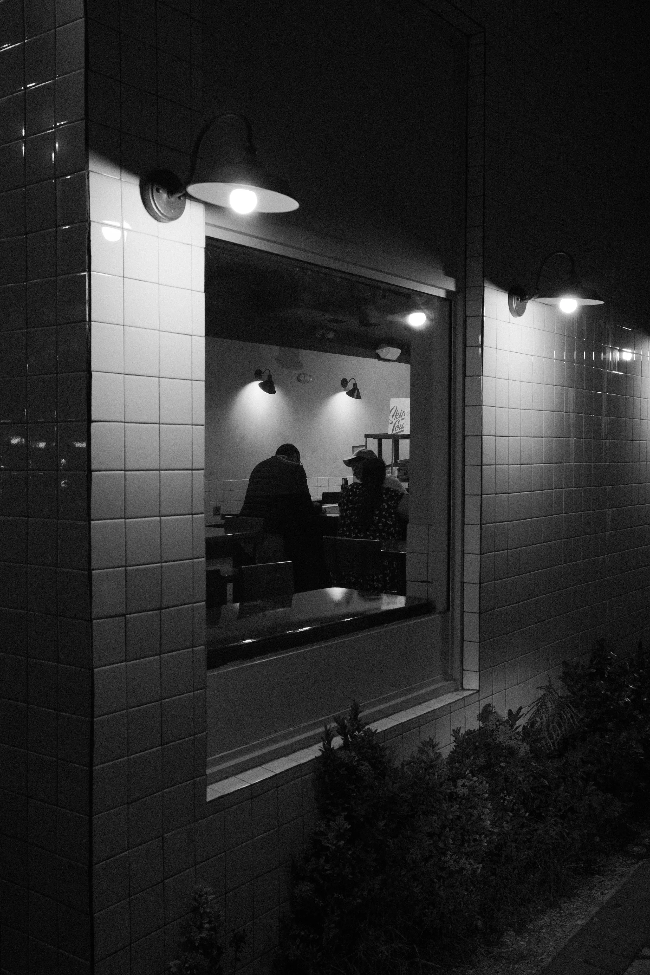 Nighttime view outside a restaurant window showing two people seated at a table inside, illuminated by interior lighting, with exterior wall-mounted lamps and bushes outside.