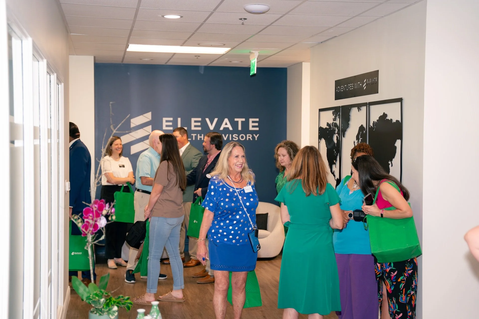 Group of people in an office lobby, chatting and smiling, with a large sign reading 'ELEVATE' on the wall behind them.