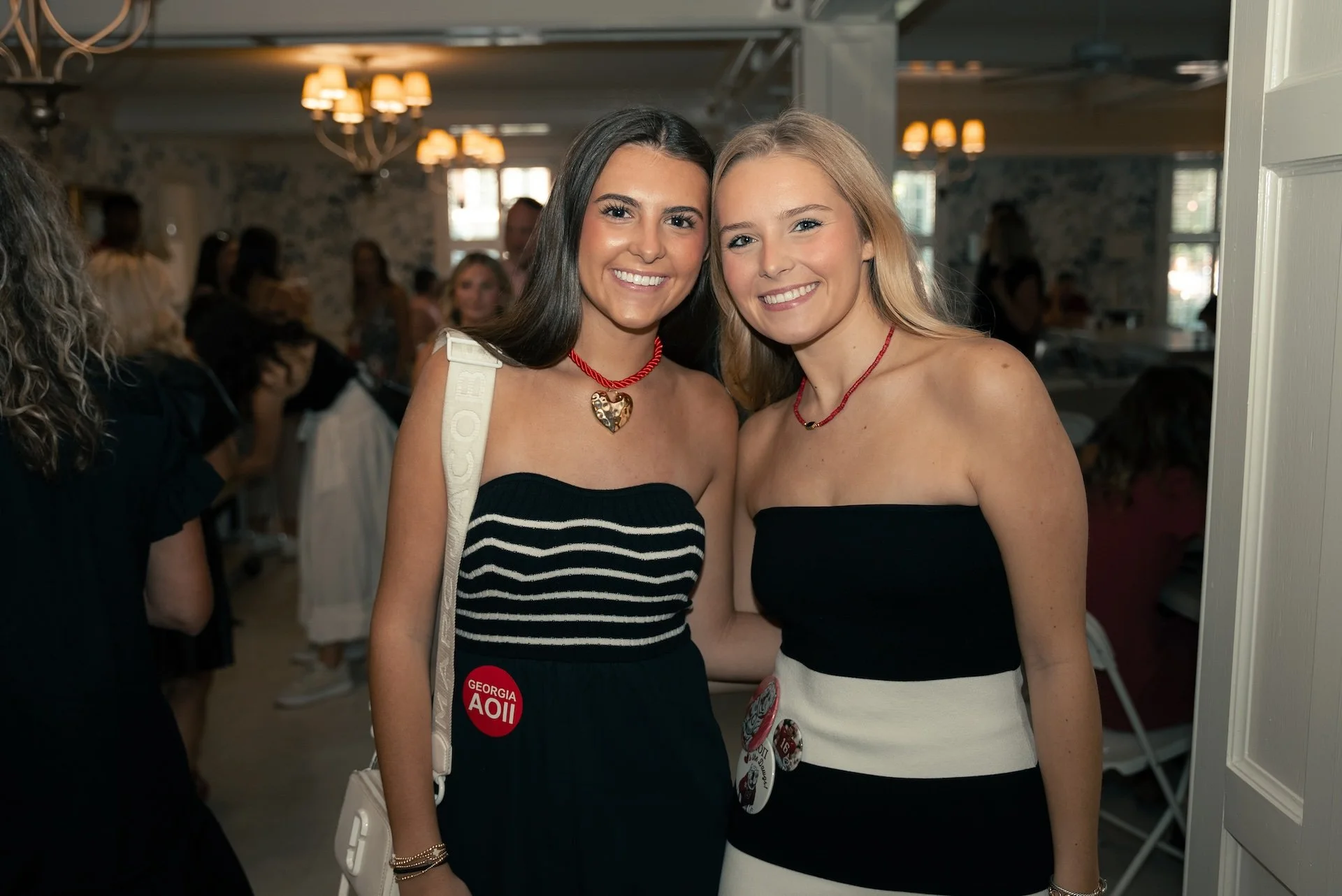 Two young women smiling and standing close together in a social gathering, wearing strapless dresses, with one carrying a white shoulder bag, in an indoor room with other people and warm lighting.