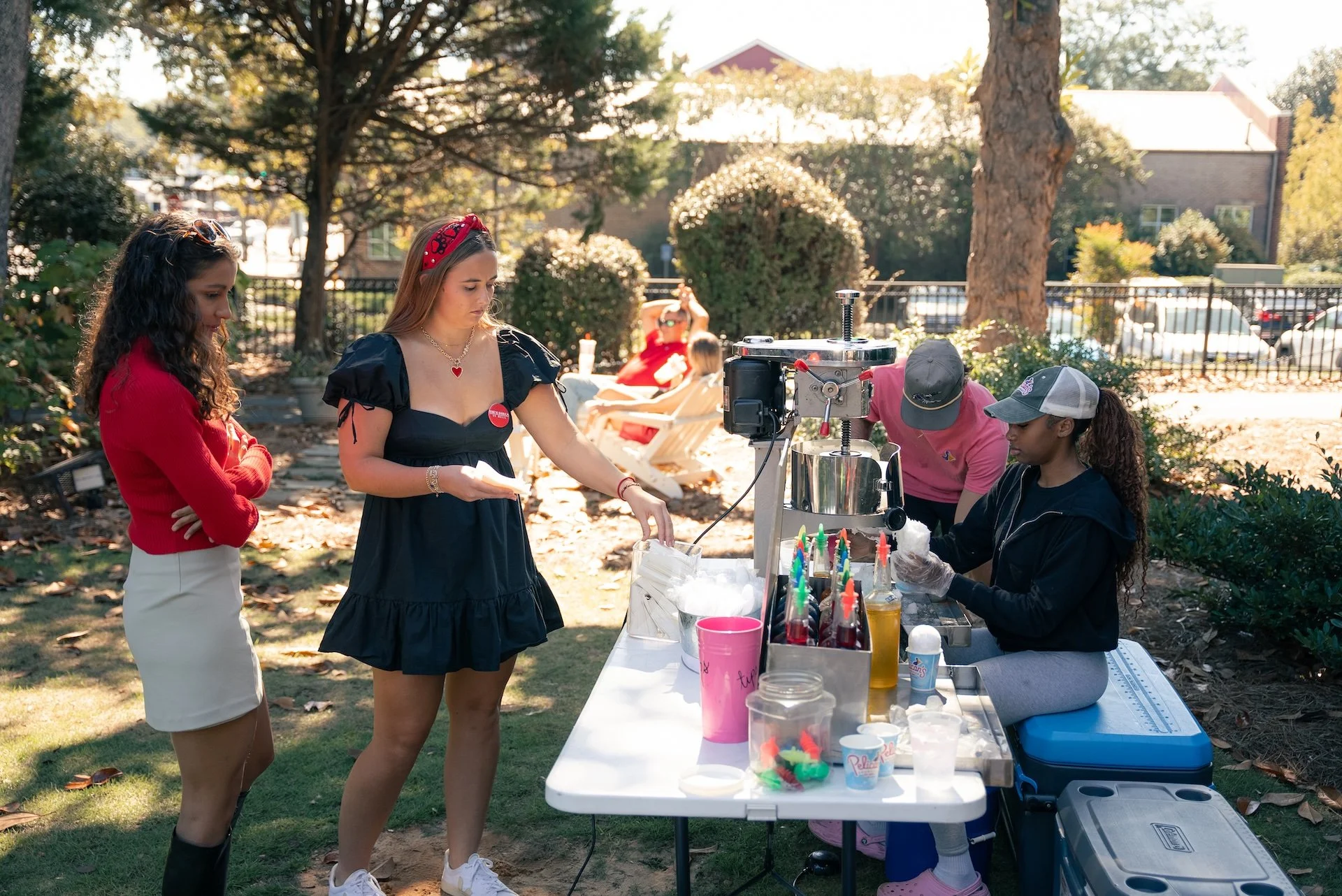 Two women and an individual serving cotton candy at a park fair stand with trees and cars in the background.