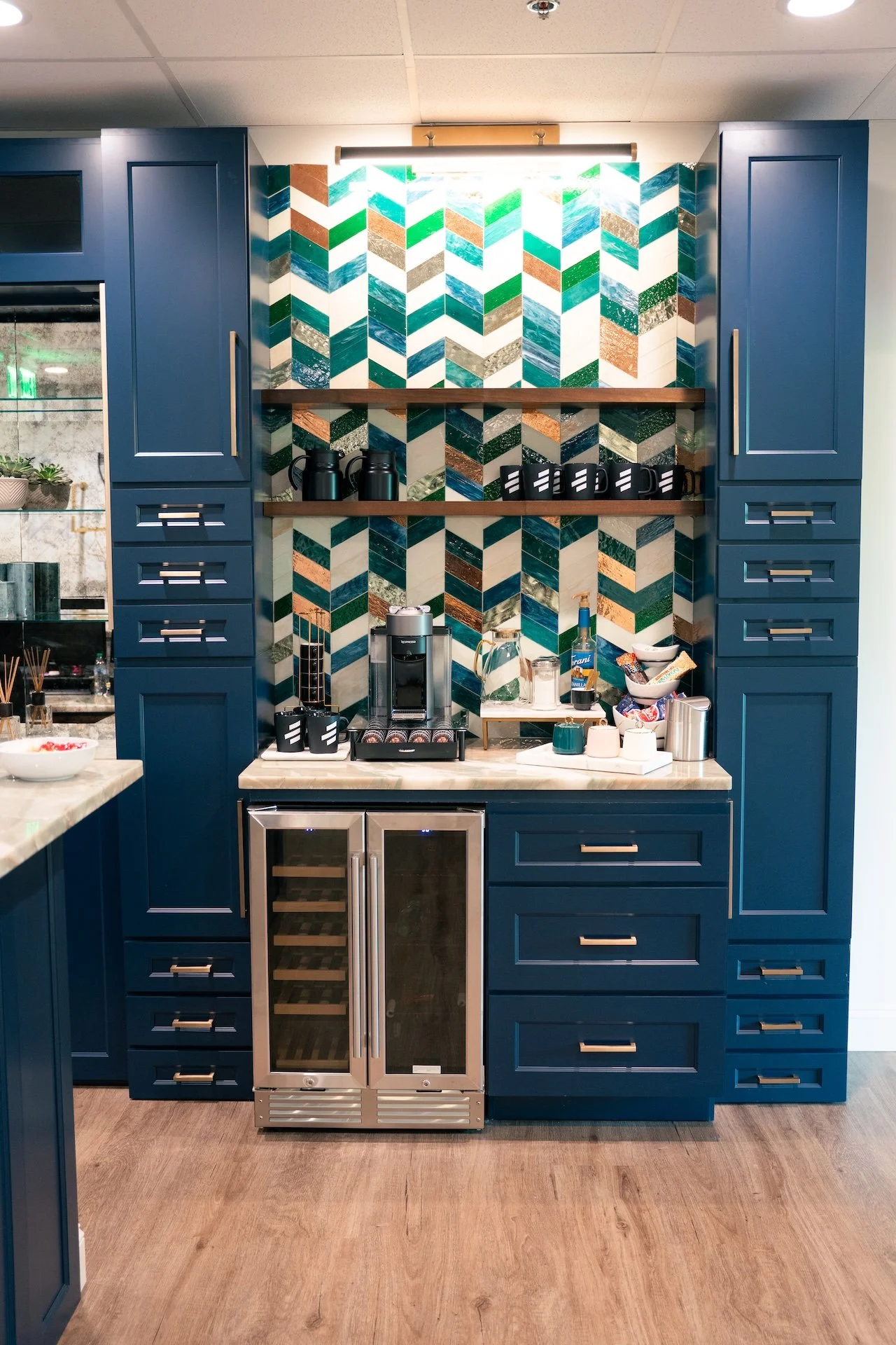 A kitchen with blue cabinets, a patterned tile backsplash in blue, green, white, and brown, a small wine cooler, and a marble countertop with coffee-making equipment and kitchen accessories.