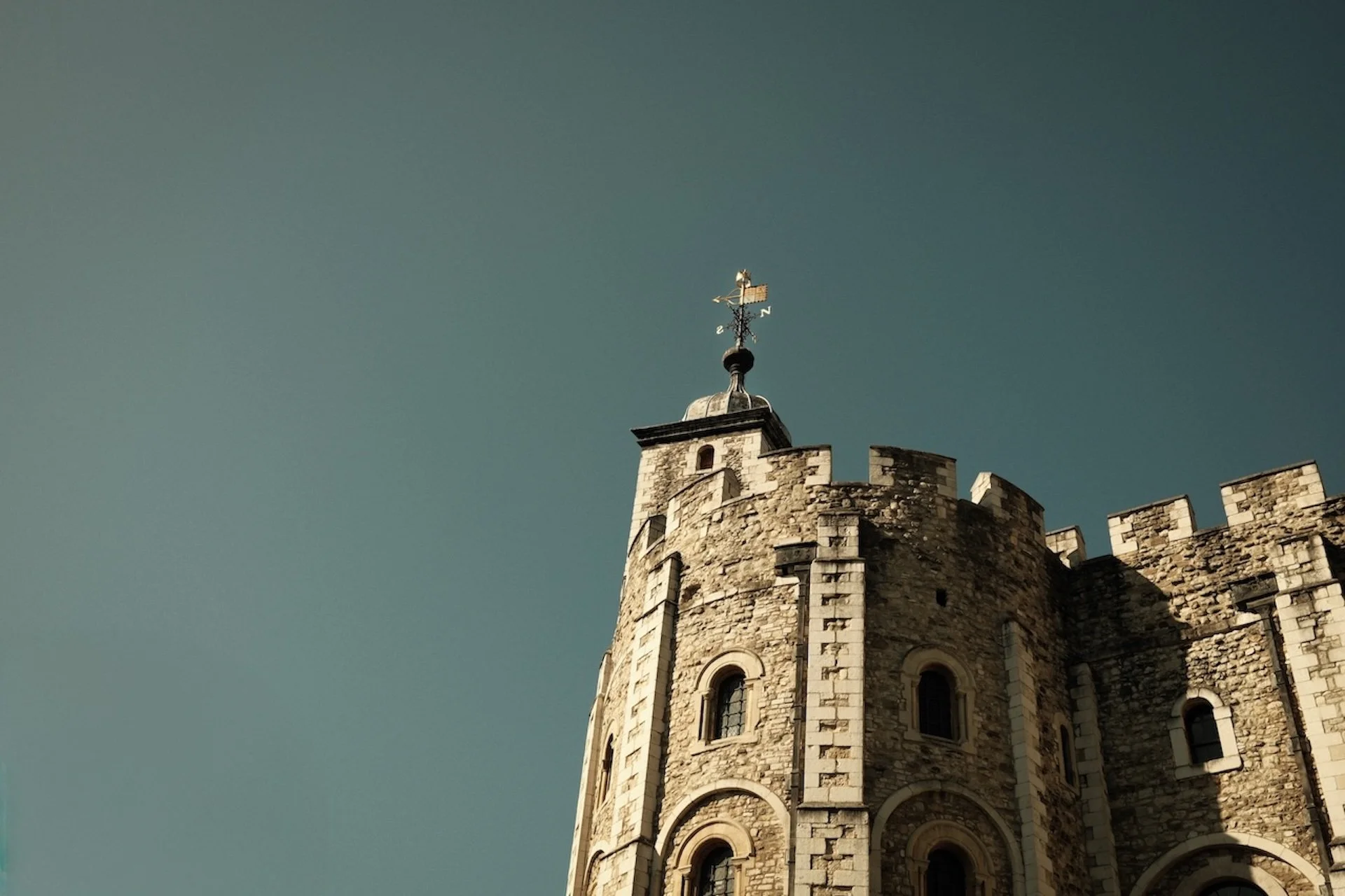 Close-up of a stone castle tower with a weather vane on top, against a clear sky.