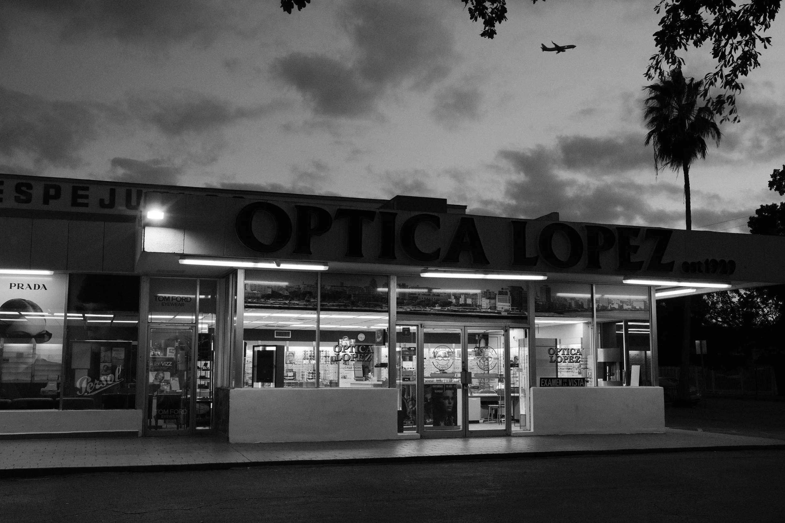 Black and white photo of an optician store named 'Optica Lopez' with illuminated signs, glass windows displaying brands like Prada and Tom Ford, and a tree and airplane in the sky above.