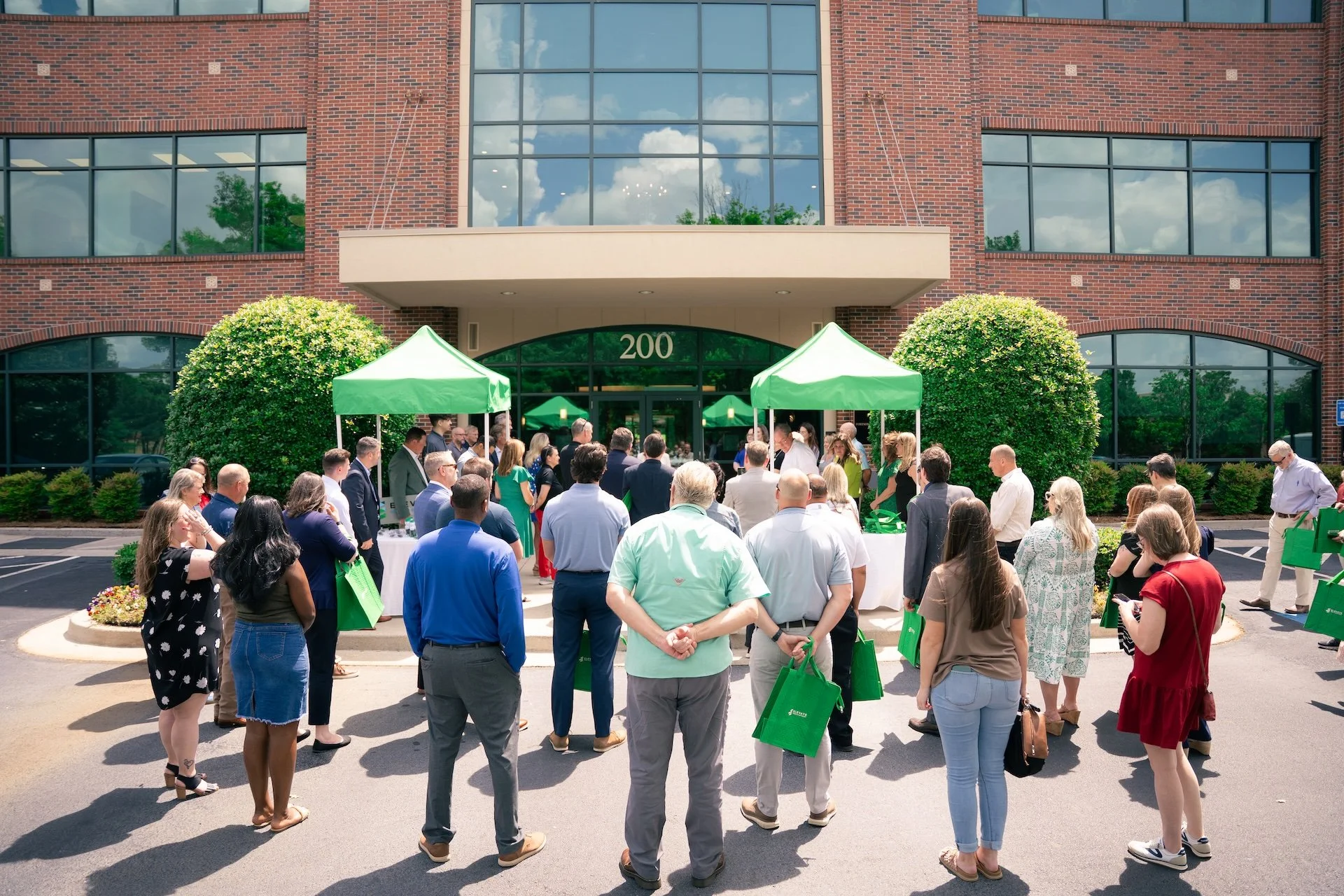 People gathered outside the entrance of a brick building with a glass facade, organized with green tents, during a daytime event.