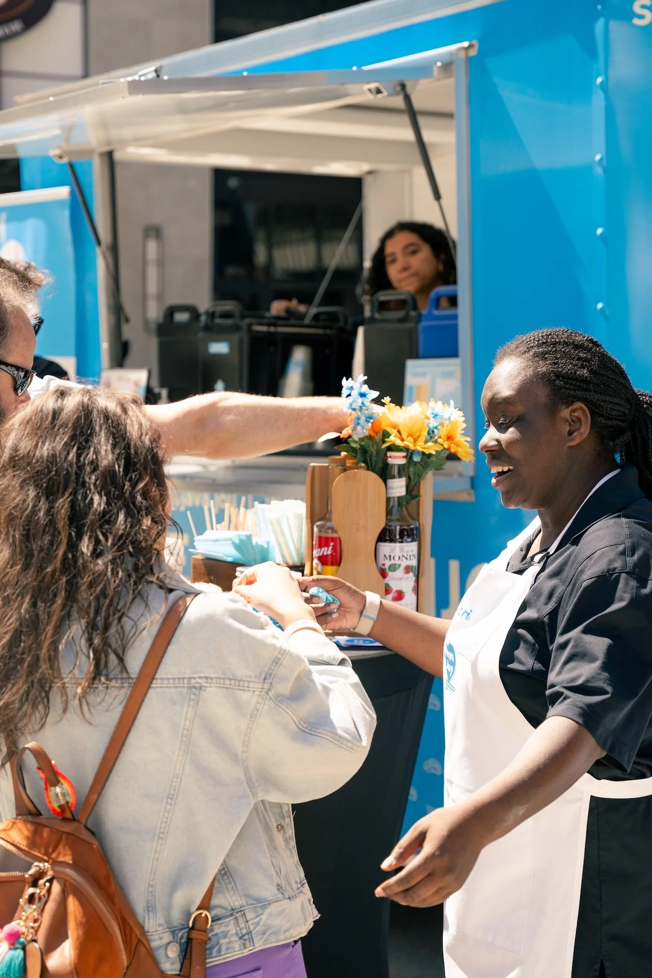 A woman in a black uniform with a white apron serves a customer at a blue food truck, with a second employee visible inside. The customer is a woman with brown curly hair and a tan denim jacket, receiving a product. A small vase with blue and yellow 