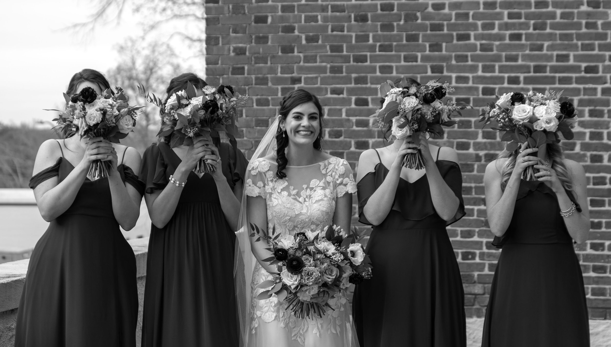 A bride in a wedding dress holding a bouquet of flowers, standing between five bridesmaids wearing dark dresses and holding bouquets in front of their faces, in front of a brick wall.