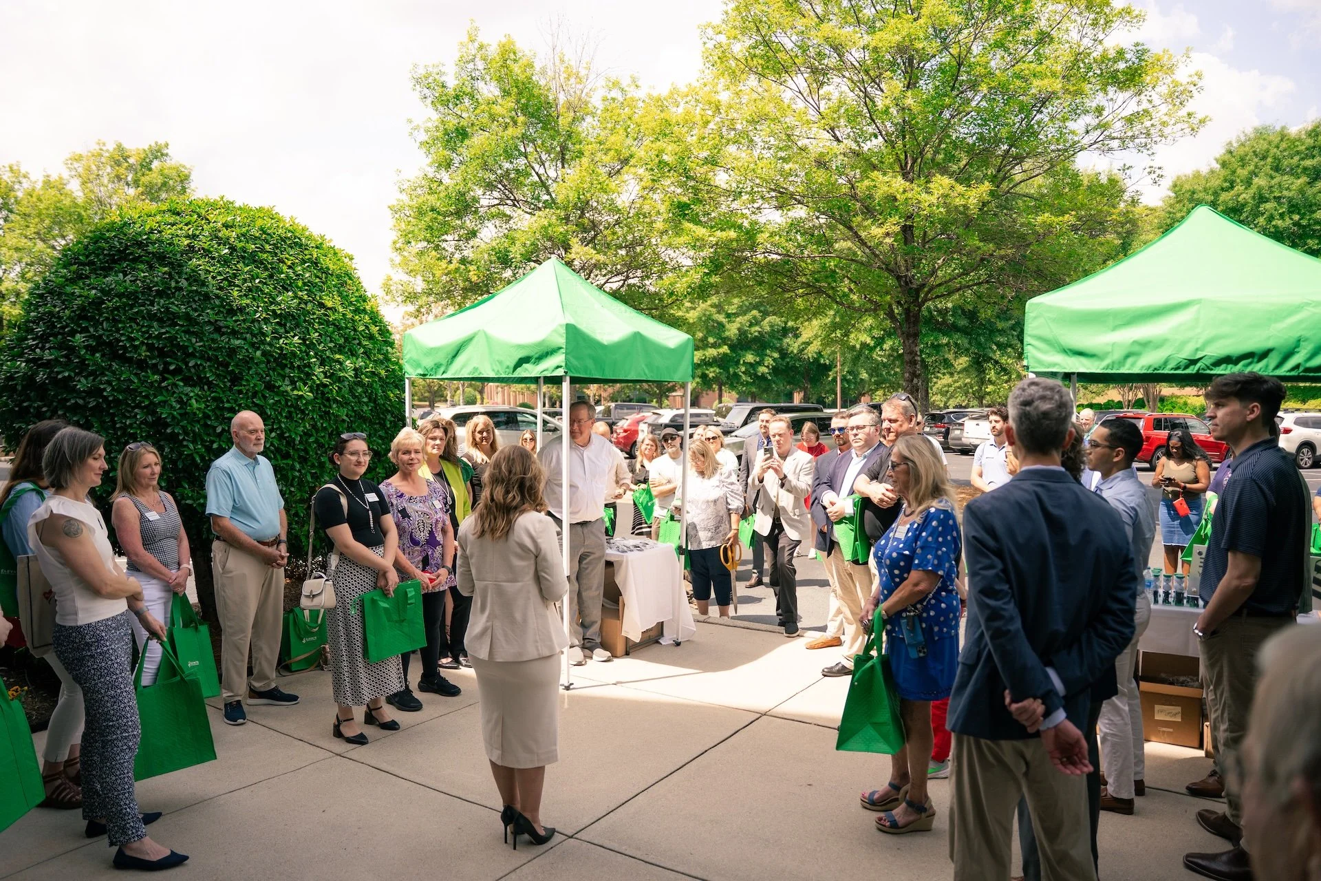 Group of people gathered outdoors under two green tents for an event, surrounded by trees and parked cars.