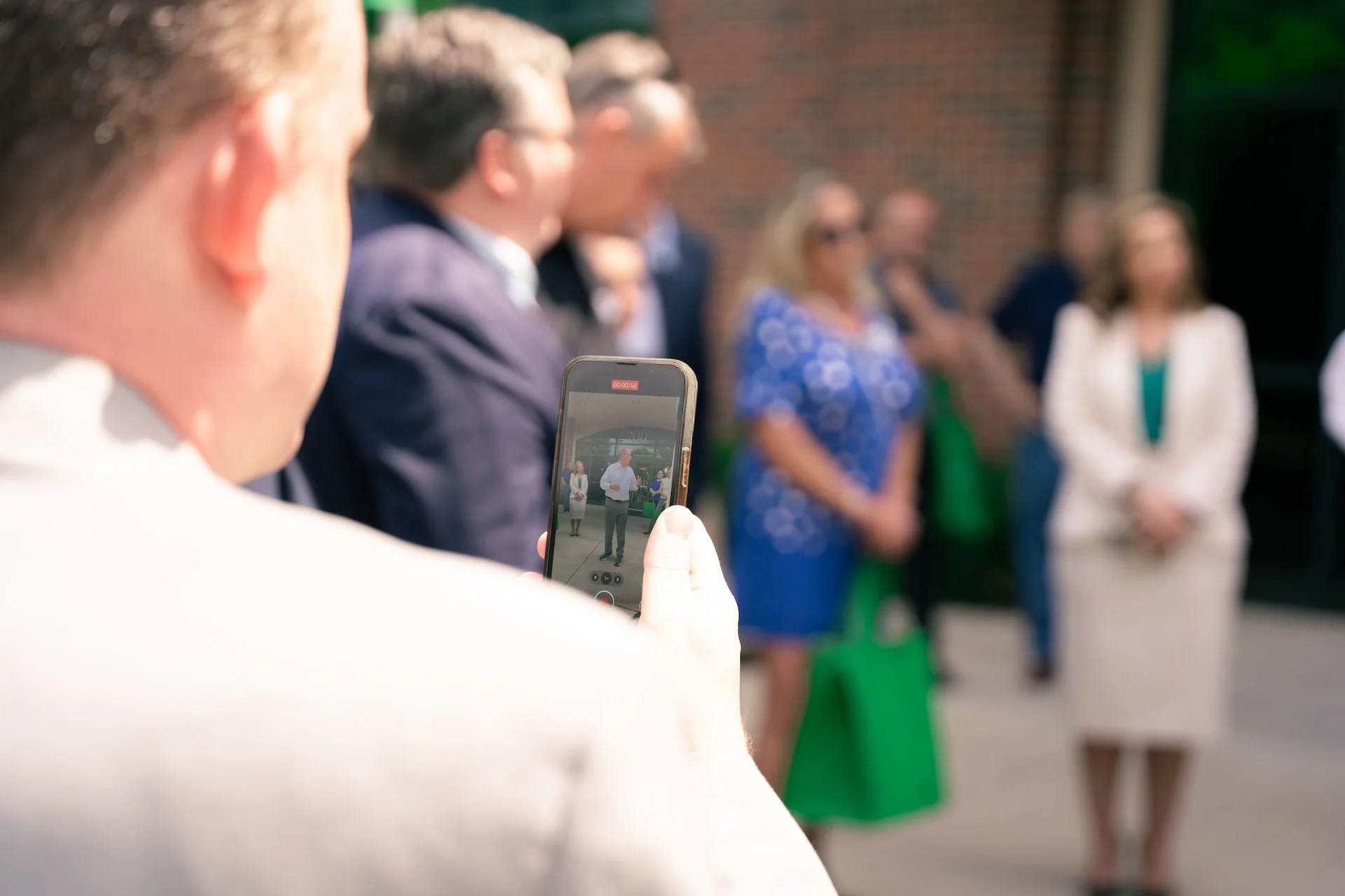 Person taking a photo with a smartphone at an outdoor event with a group of people standing in a row.