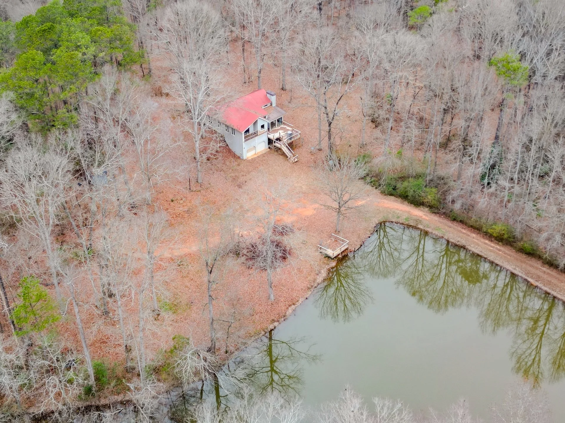 An aerial view of a house with a red roof next to a small pond, surrounded by leafless trees in a wooded area.