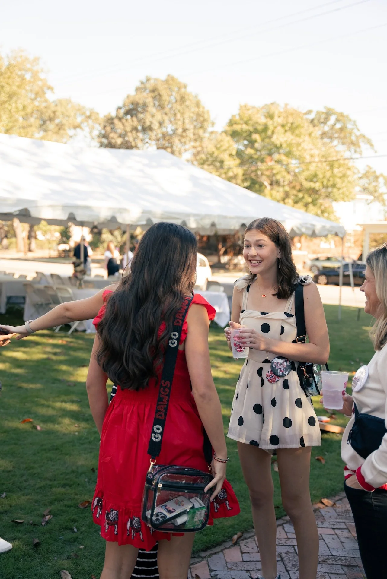 Three women are standing outdoors and talking. They are holding drinks and smiling. Behind them, there is a white tent and trees, suggesting a social gathering or event.