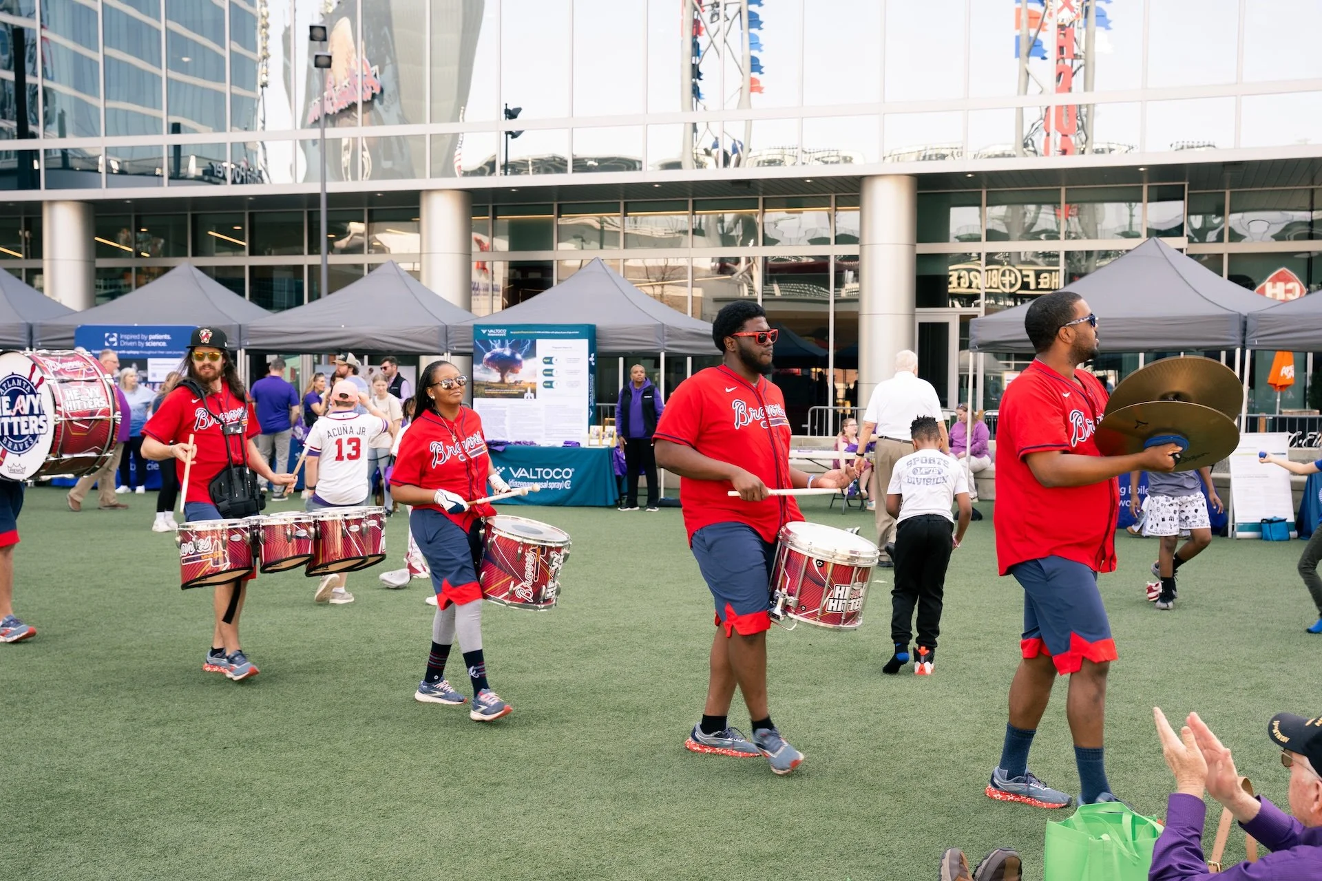 A group of four people wearing red shirts and shorts playing drums and cymbals in a parade on a grassy field outside a large modern building with glass windows and tents in the background.