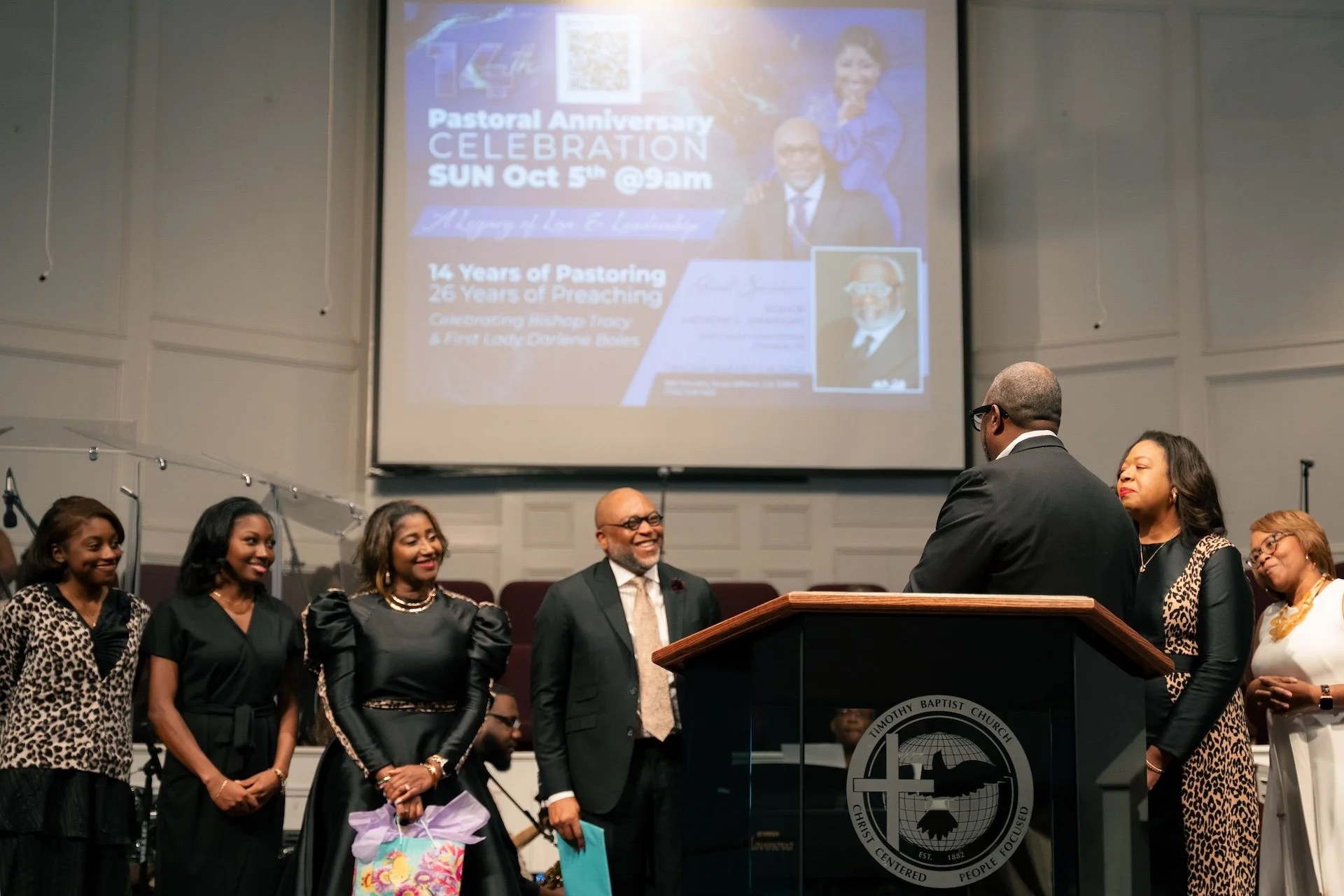 A group of people standing on stage in front of a podium and a large screen displaying a flyer about a pastoral anniversary celebration. The group includes four women and two men, some in formal and semi-formal attire, with smiling expressions. The s