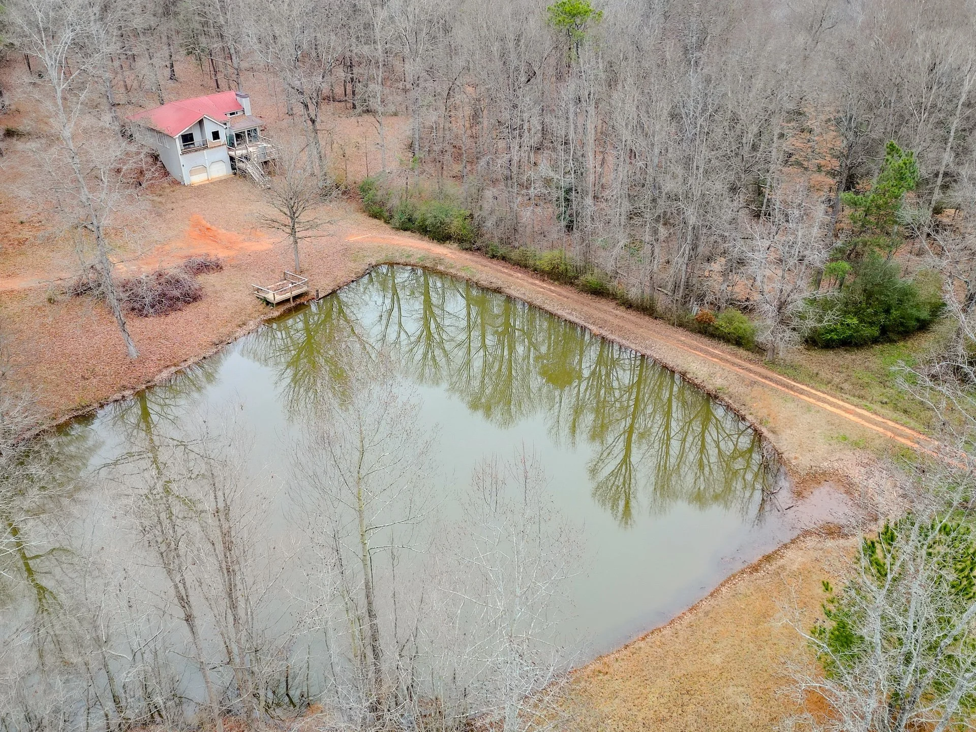 A house near a pond surrounded by trees with mostly bare branches, in a rural setting during late fall or winter.