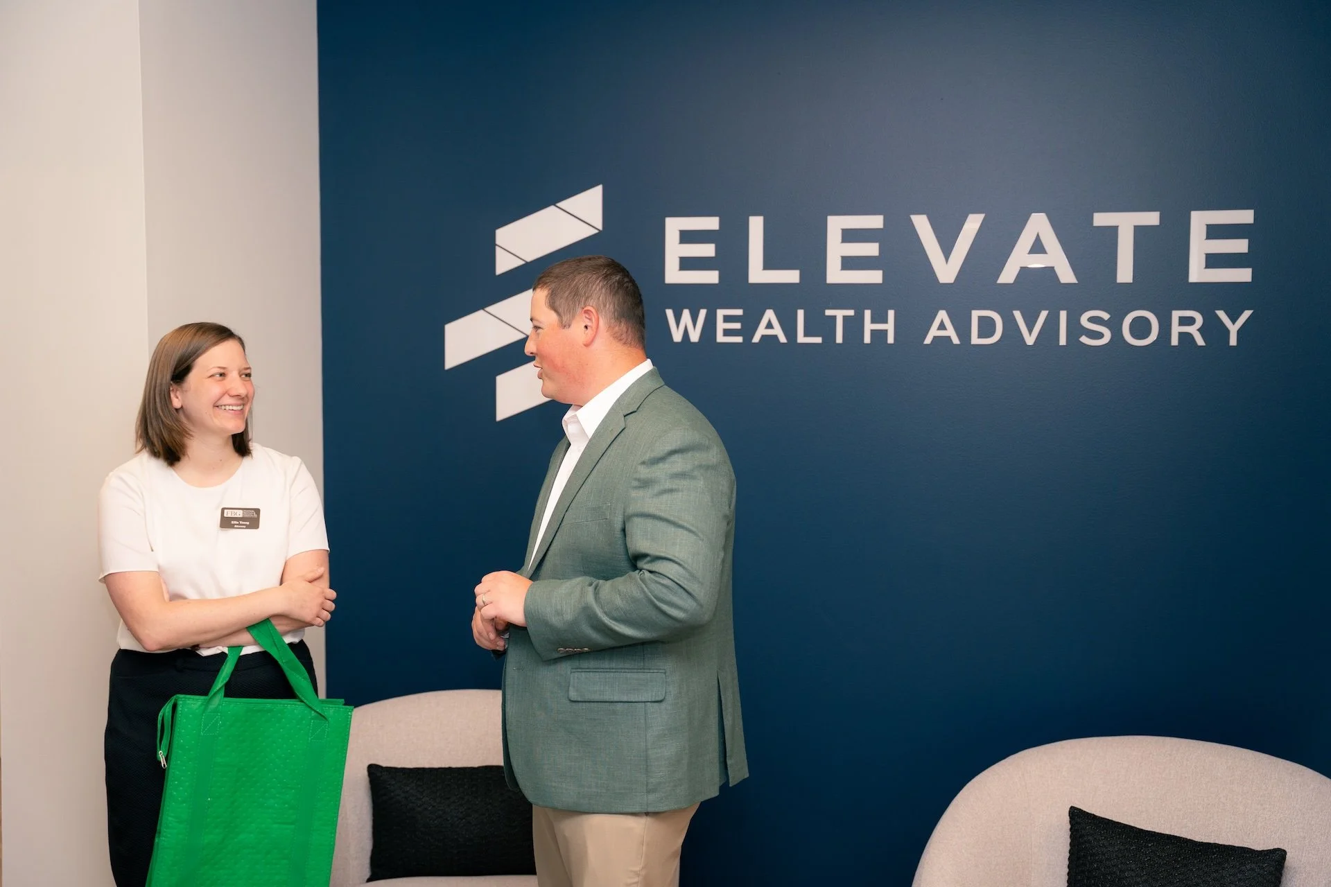 A woman and a man are talking in an office lobby with a sign that says "ELEVATE WEALTH ADVISORY." The woman is smiling, holding a green tote bag, and wearing a white shirt with a name tag. The man is dressed in a light gray blazer and beige pants.