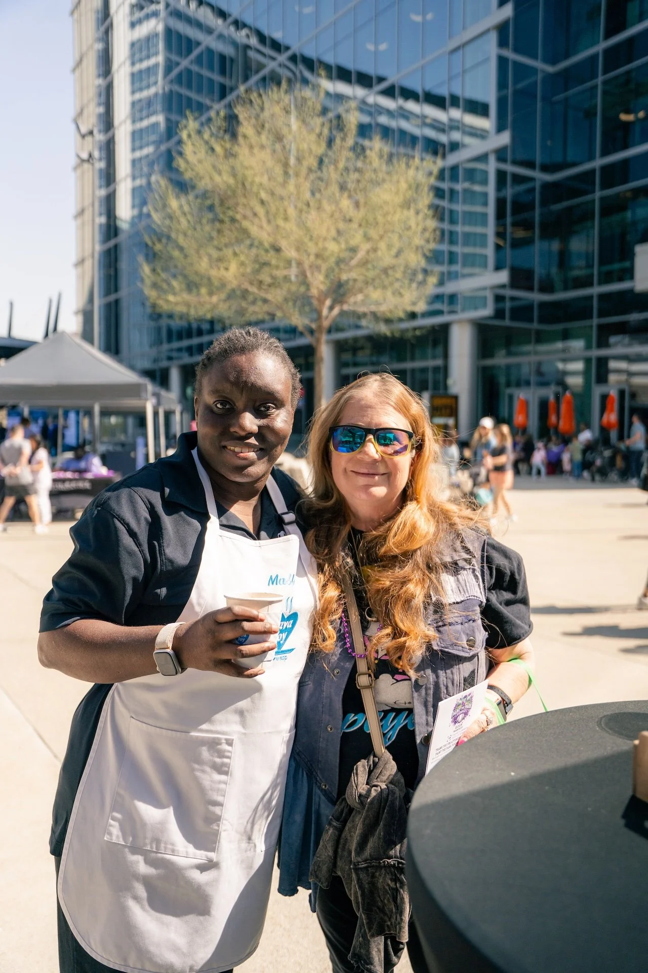 Two women standing outdoors at an event, smiling at the camera. One woman is holding a cup, wearing a white apron, and the other woman is wearing sunglasses and a black shirt. In the background, there are tents, chairs, and a modern glass building.