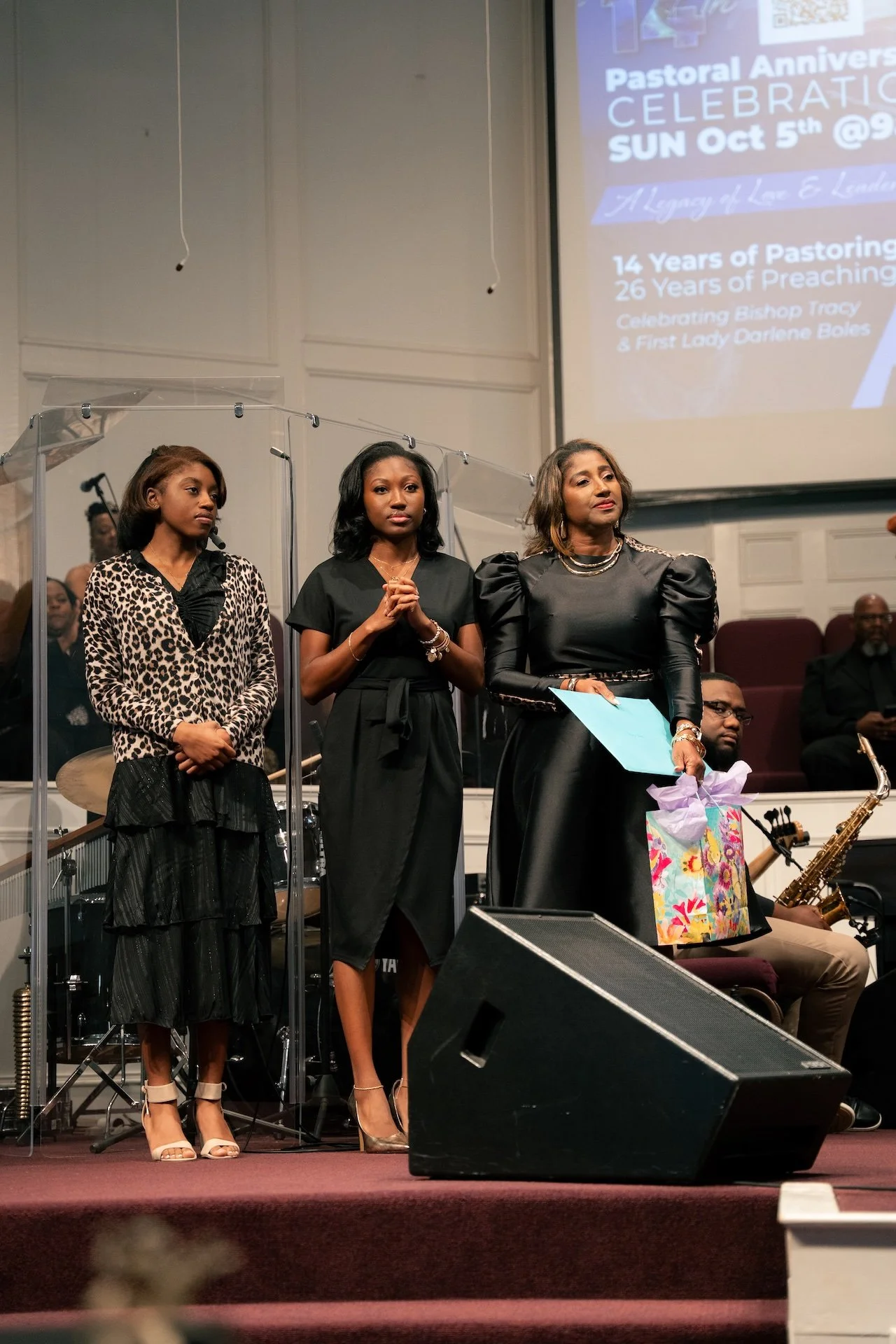 Three women standing on stage during a church anniversary celebration, with a large screen behind them displaying event details and clergy members seated in the background.