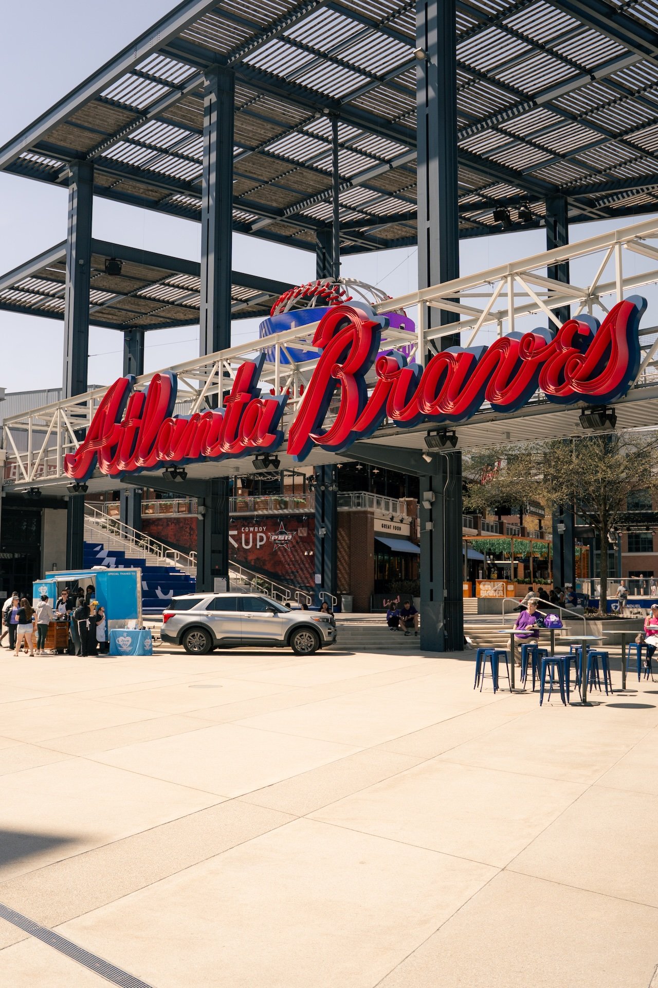 The entrance to Atlanta Braves stadium featuring a large, red neon sign that says 'Atlanta Braves' with a baseball sculpture above it. There are people, cars, and outdoor seating visible in the foreground.