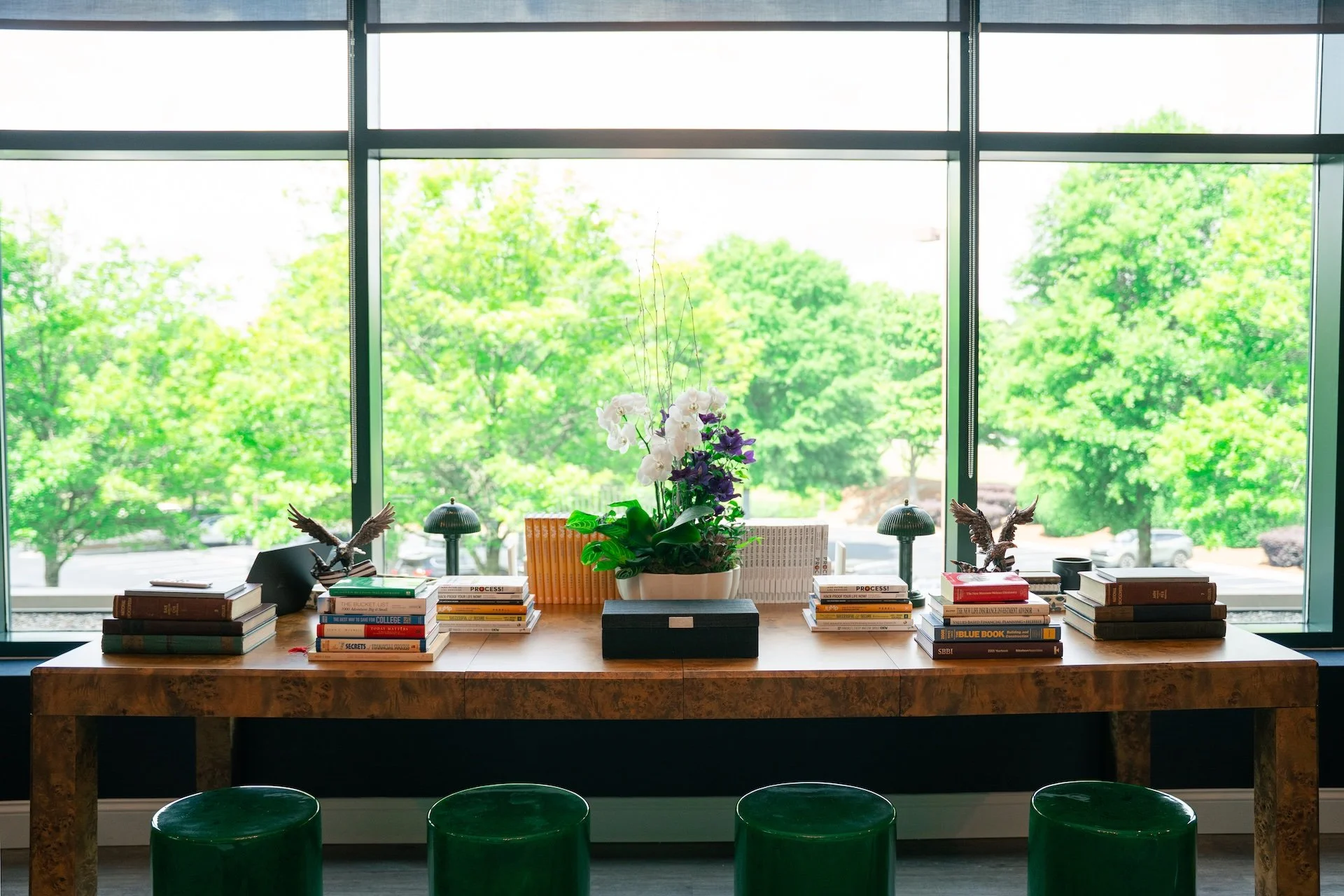 A decorative office or conference room with a wooden table and green stools, a potted orchid in the center, books and figurines on the table, and large windows with a view of green trees outside.