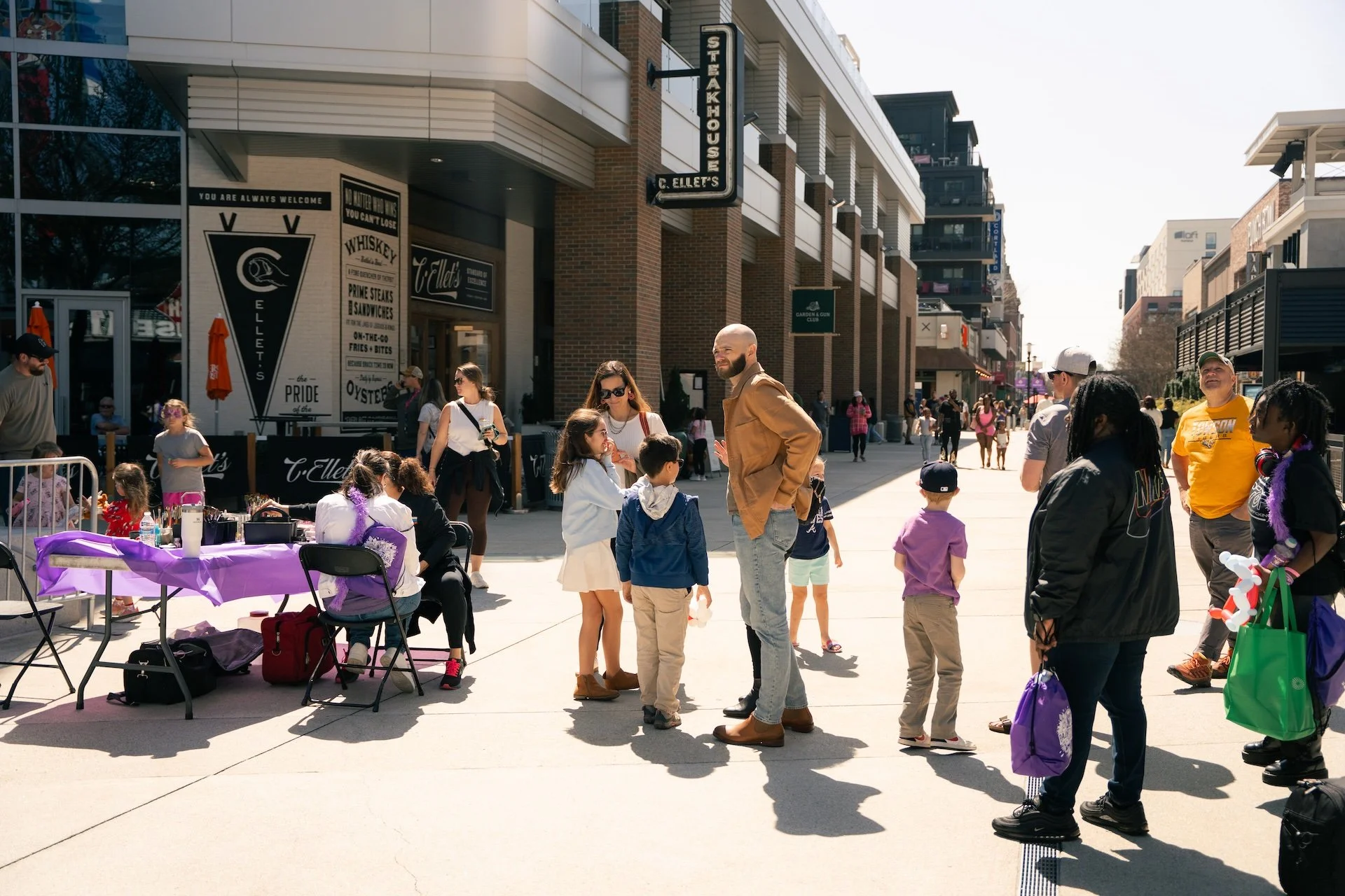 People gather outside a restaurant with signage reading 'C. Ellet's' on a city street during the day. Some people are seated at tables, while others are standing and talking. The scene includes children and adults, with some carrying shopping bags an