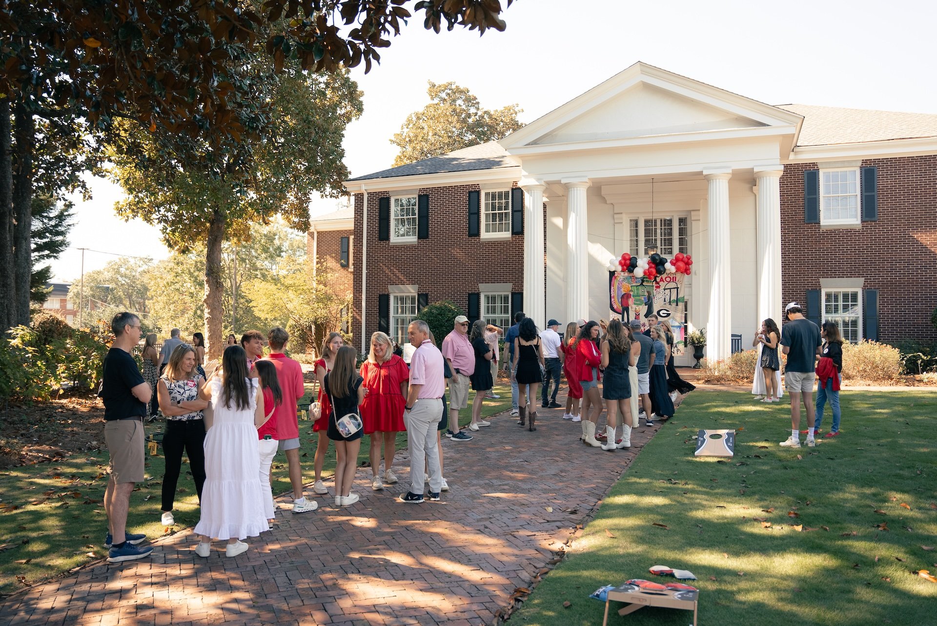 A group of people gathered outside a brick house with a stoop, during daytime. The scene includes children and adults, some in casual clothing, standing on a brick pathway and grass lawn, with trees and a sunny sky in the background. There are decora
