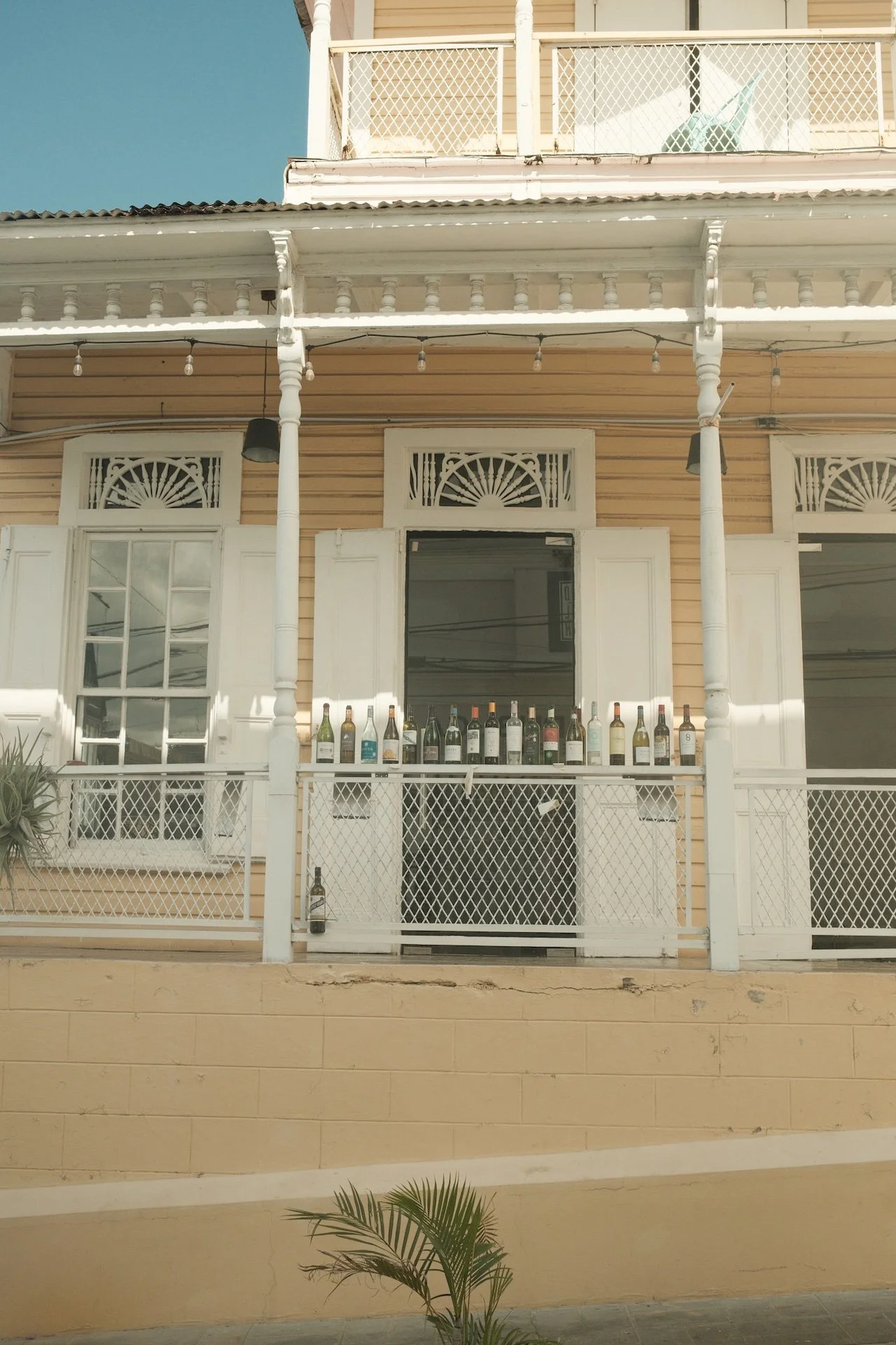 Exterior of a yellow house with white trim, featuring a porch with railings and a row of various bottles lined up along the railing.
