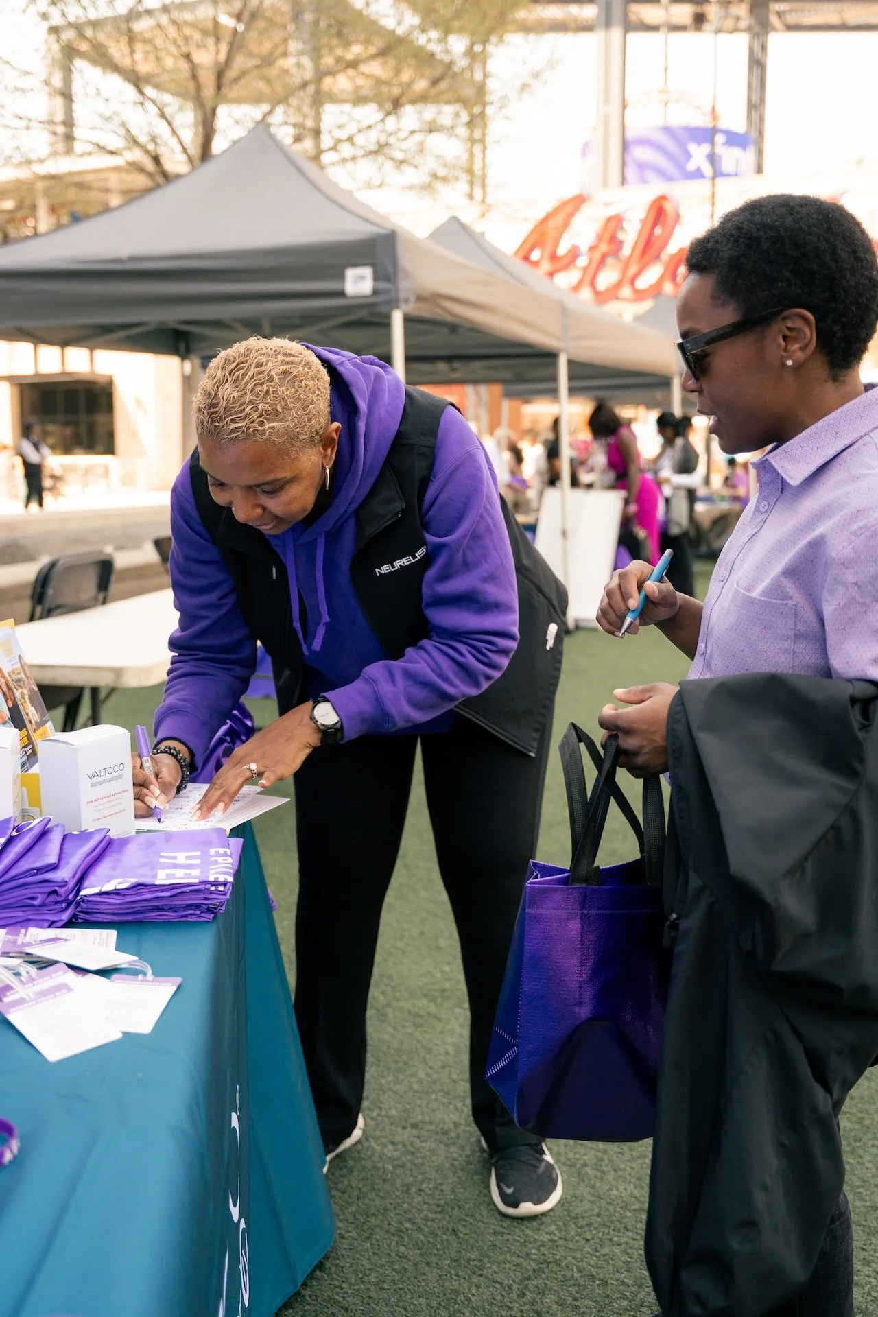 Two women at an outdoor event, one signing a paper at a table with purple lanyards and informational materials, while the other stands next to her holding a purple tote bag and a pen, with tents and people in the background.