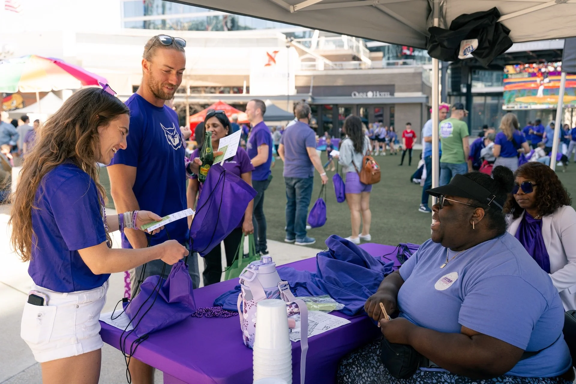 People at a registration table at an outdoor event, wearing matching purple shirts, with others in the background under tents and umbrellas.