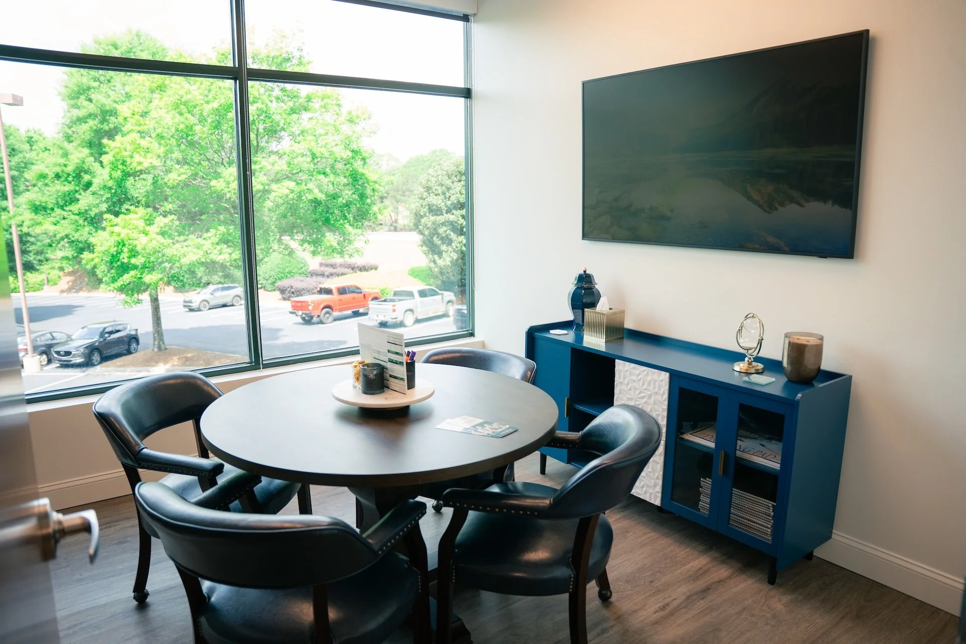 A modern conference room with a round table and five black chairs. Large window with a view of green trees and parked cars. Blue sideboard with decorative items and a large flat-screen TV on the wall.