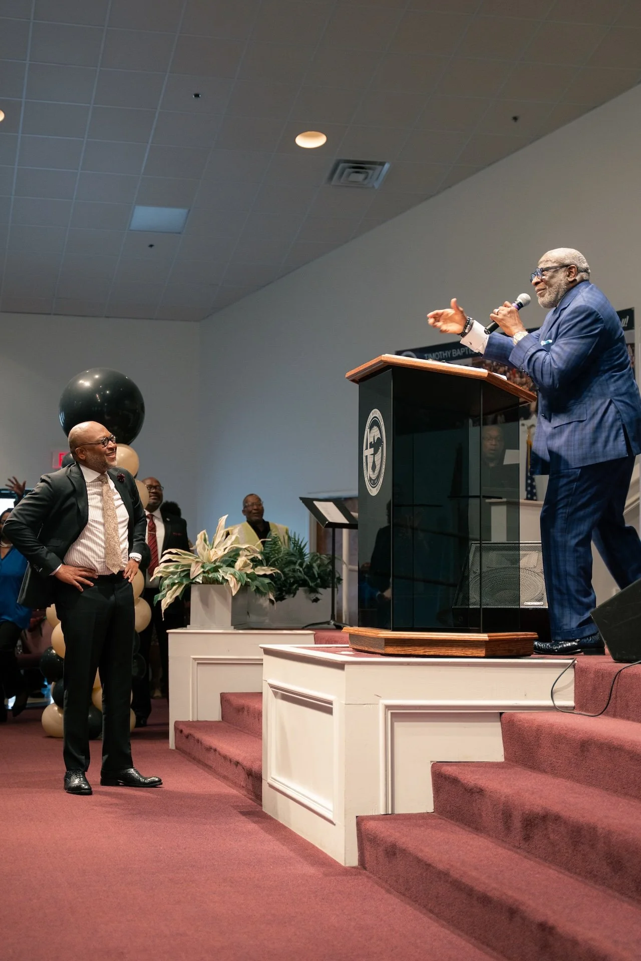 A man in a blue suit speaking at a podium on stage while another man in a dark suit and glasses listens and smiles. Several people are in the background, some with hands raised, and there are decorations including balloons and plants.