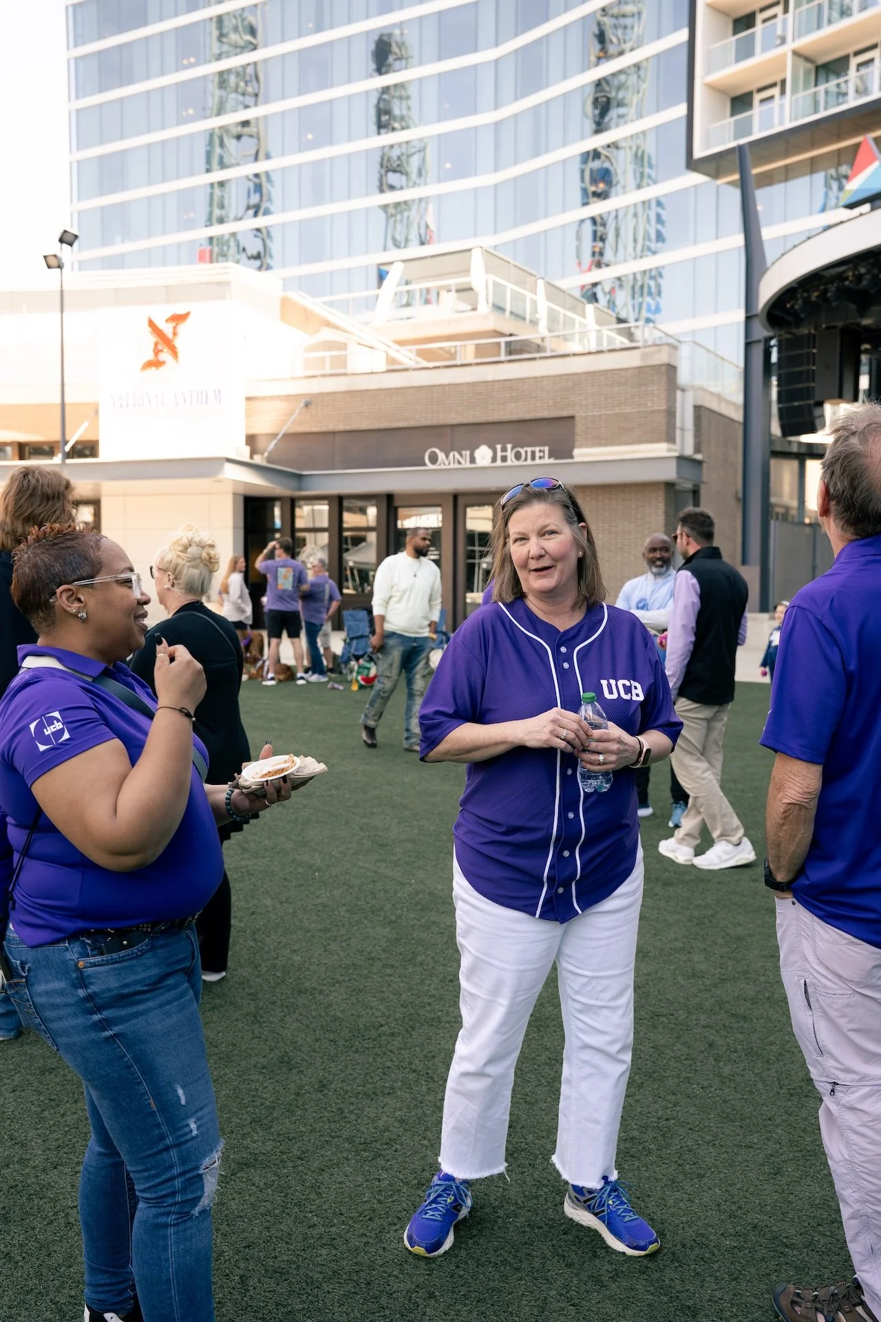 Group of people wearing purple UCB shirts socializing outdoors in front of the Omni Hotel.