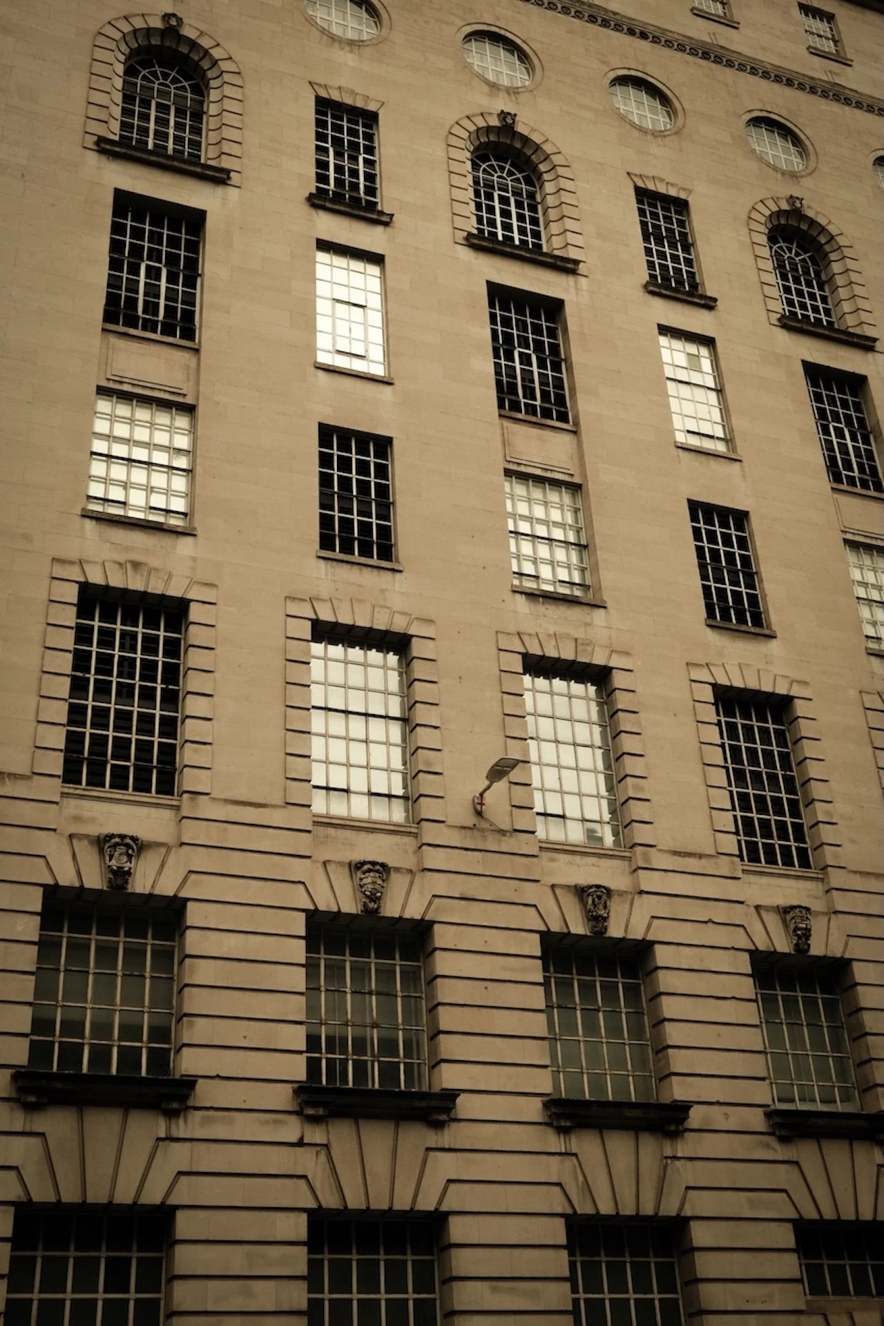 A tall beige city building with multiple windows, some with metal bars, and decorative architectural details near the window frames. There's a street lamp attached to the building.