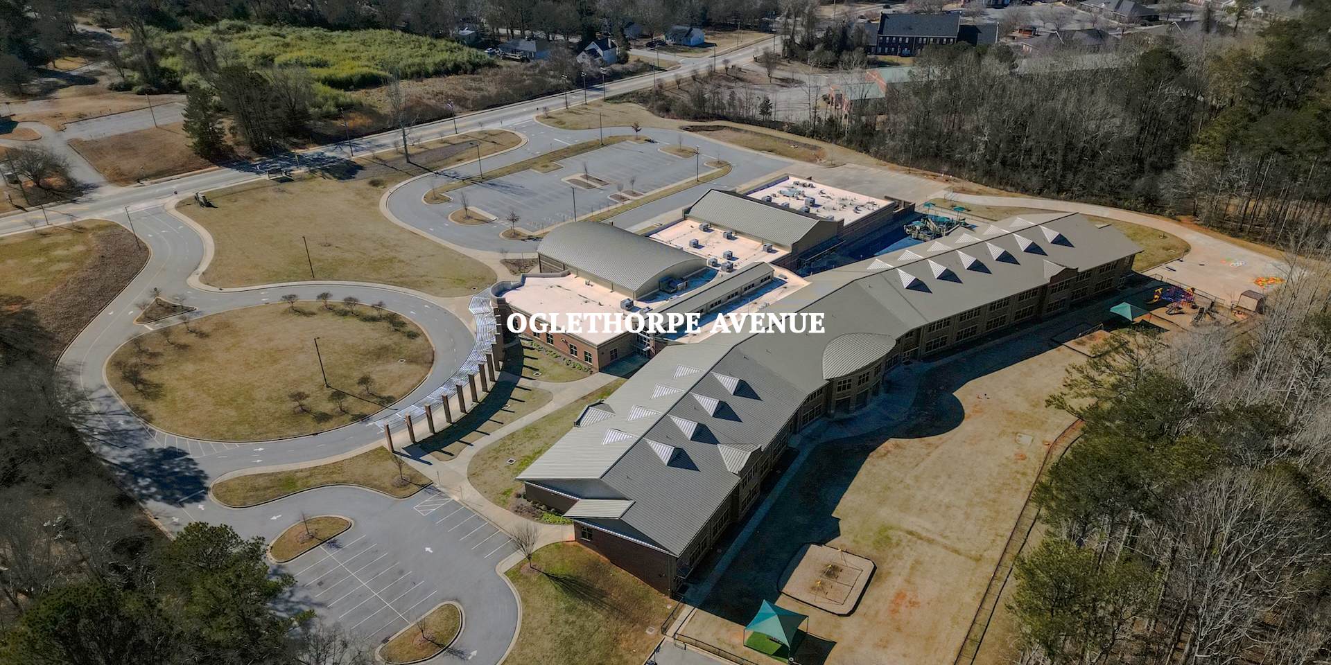 An aerial view of a large school building with parking lots, a playground, and surrounding trees on Oglethorpe Avenue.