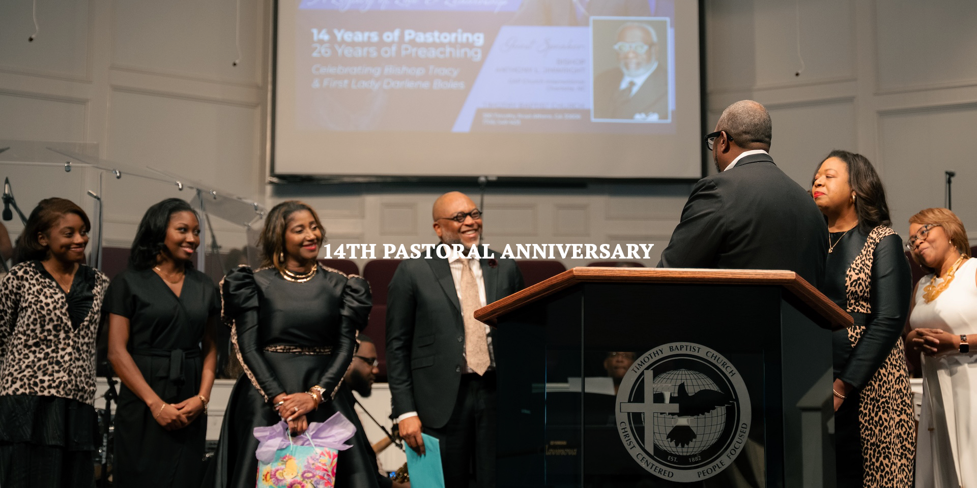 A group of people standing on stage during a church celebration with a podium that has the Timothy Baptist Church logo, a large screen in the background displaying information about the event, and seven women and one man dressed in formal attire, some wearing animal print clothing, celebrating the church's 14th pastoral anniversary.