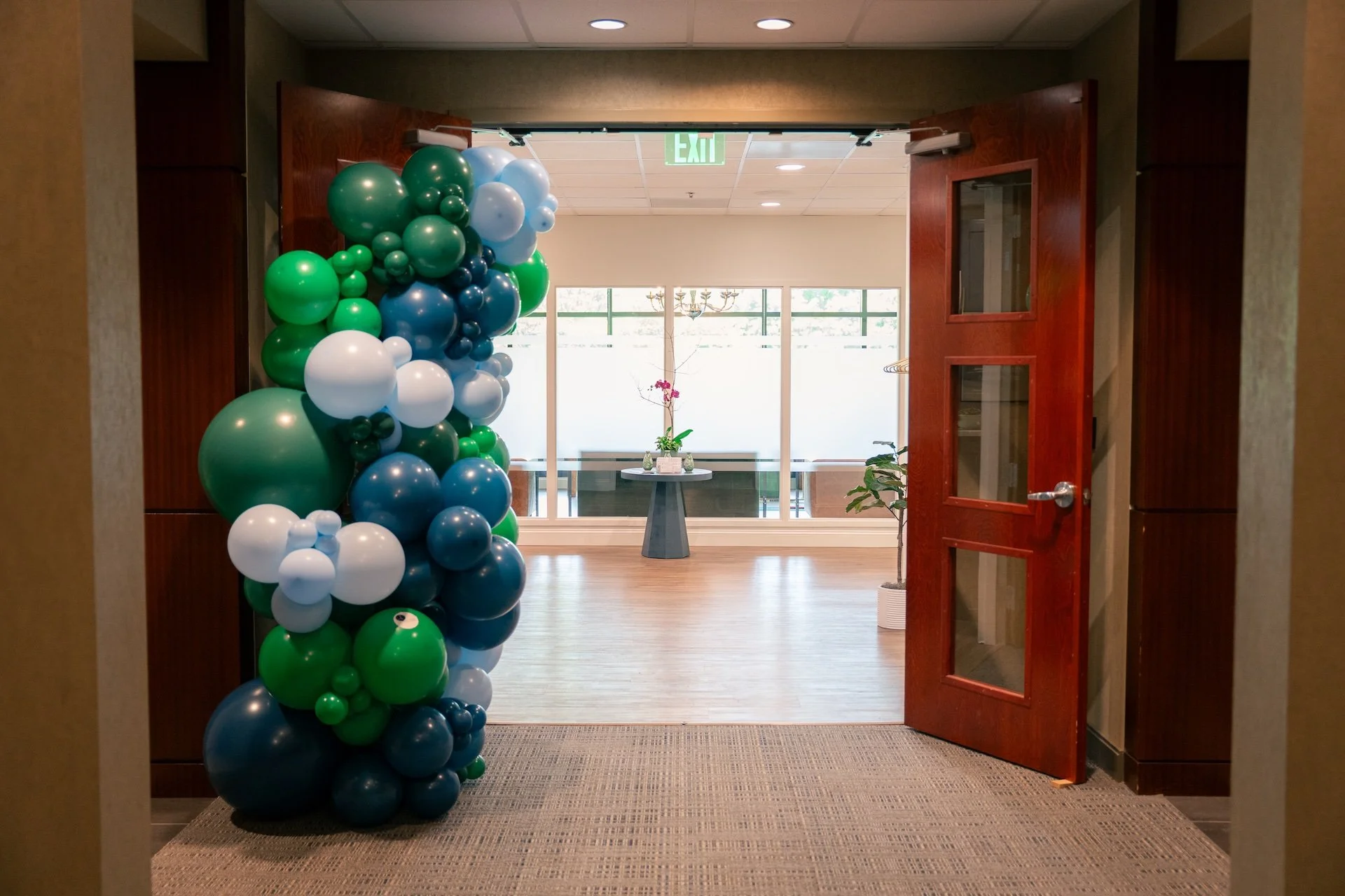 A doorway decorated with clusters of green, blue, and white balloons opening into a room with a table, potted plants, and large windows with an exit sign above.