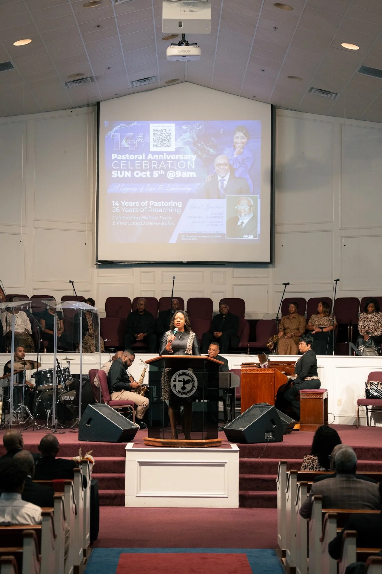 A woman speaking at a church podium during a celebration event, with a band and choir behind her, and a large screen displaying event details in the background.