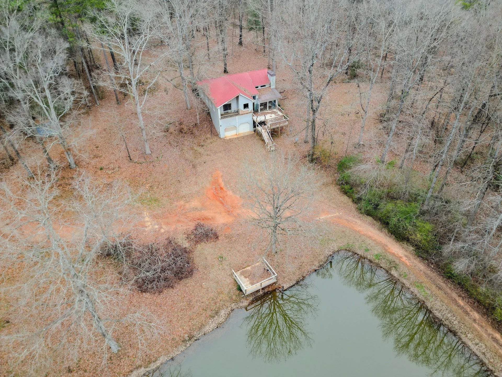 Aerial view of a house surrounded by trees near a pond, with a red roof, a deck, and a dirt driveway.